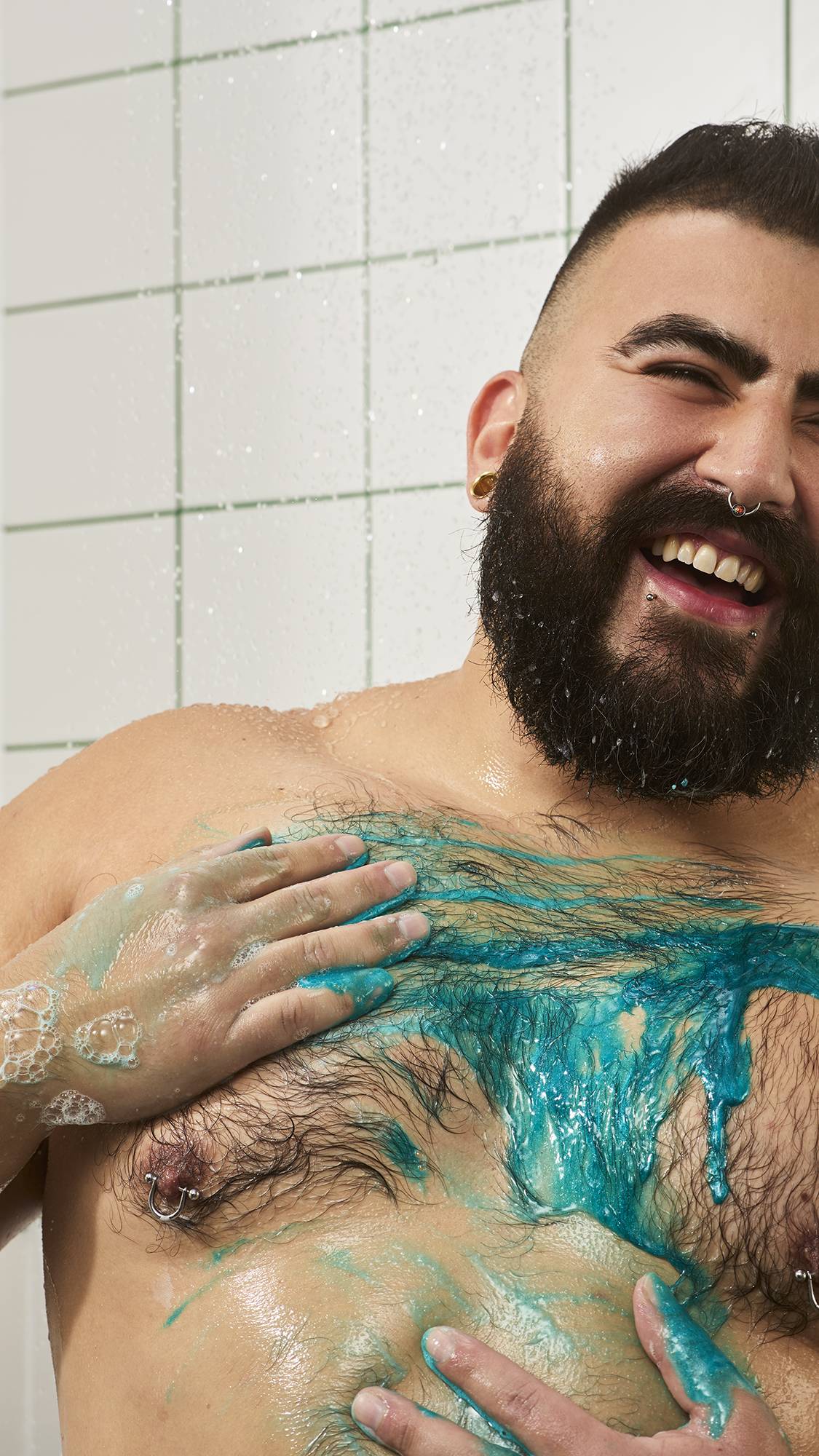 A smiling, bearded model washes in the shower, lathering vibrant shimmering blue shower gel across their chest and torso against a background of white tiles.