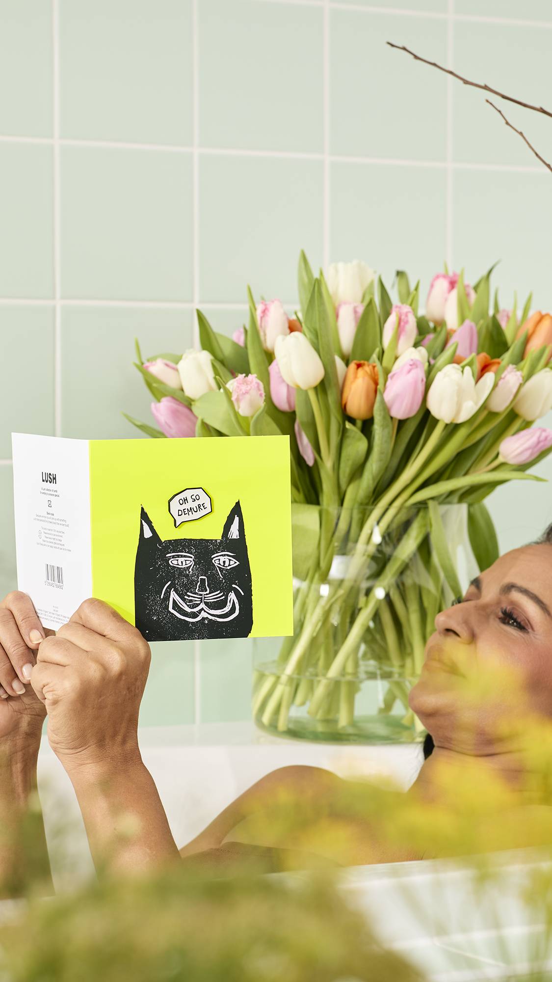 The image shows the model over the edge of a bath, holding a green Oh So Demure card featuring a black cat illustration and a speech bubble reading ‘OH SO DEMURE’. The background shows pale green tiles and a bouquet of pink and white tulips.