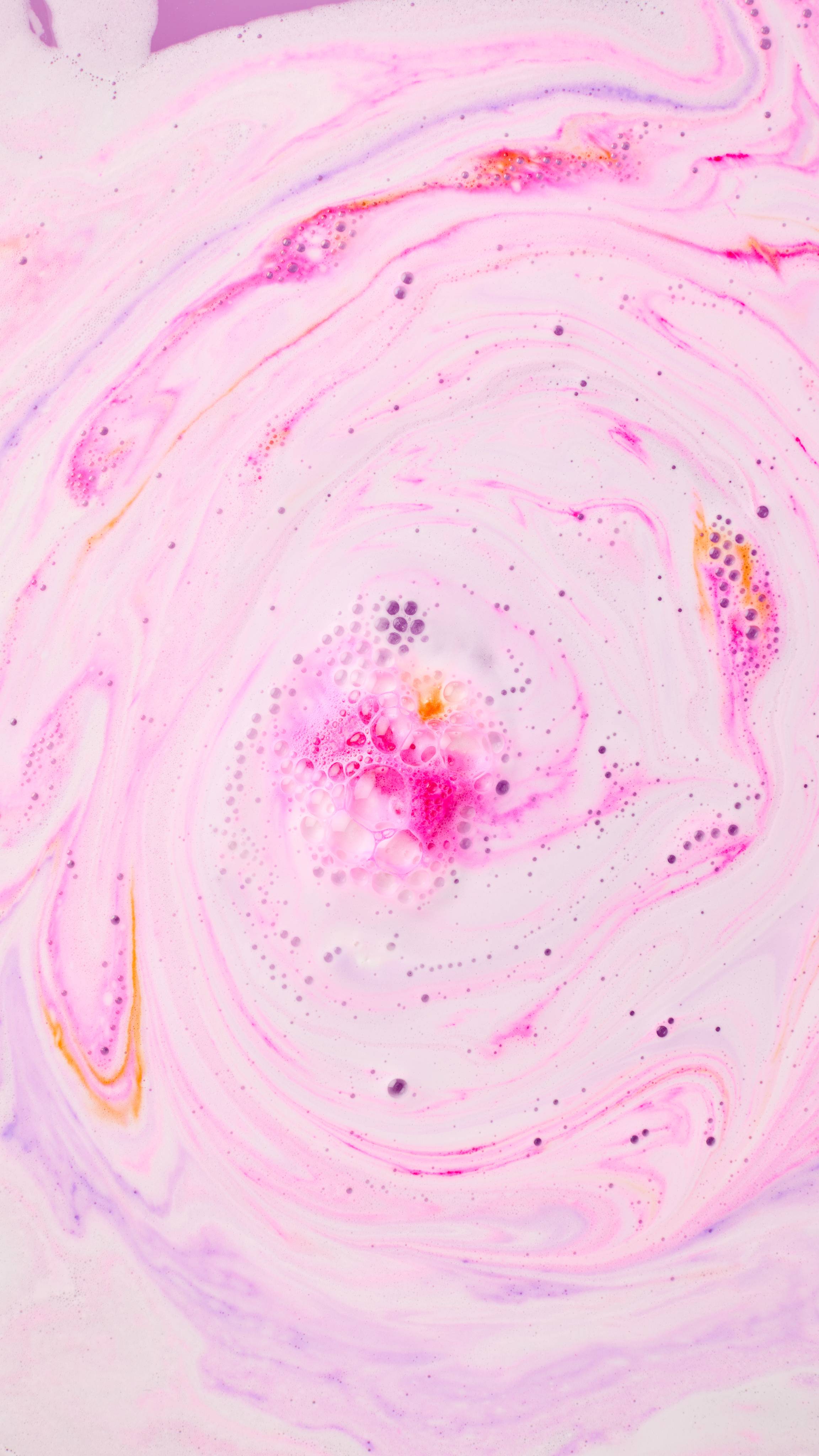 Swirling patterns of pastel pink, lilac, and white bath foam surround a central pool of vibrant pink fizz with small orange specks and glossy bubbles.