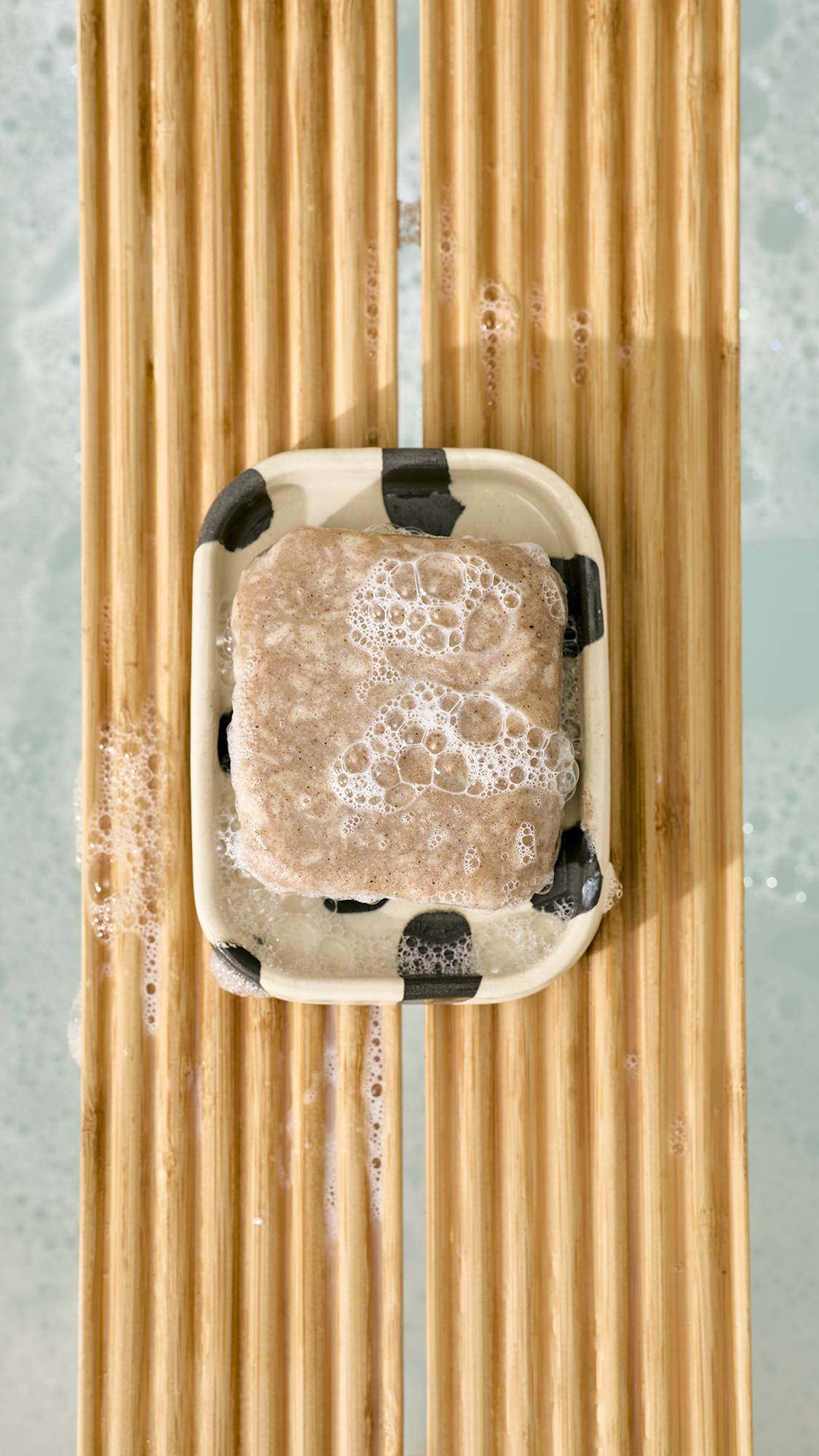 A top-down view of a bar of soapy, textured bar soap resting on a ceramic cow-print soap dish, placed on a slatted wooden bath caddy over foamy bathwater.   