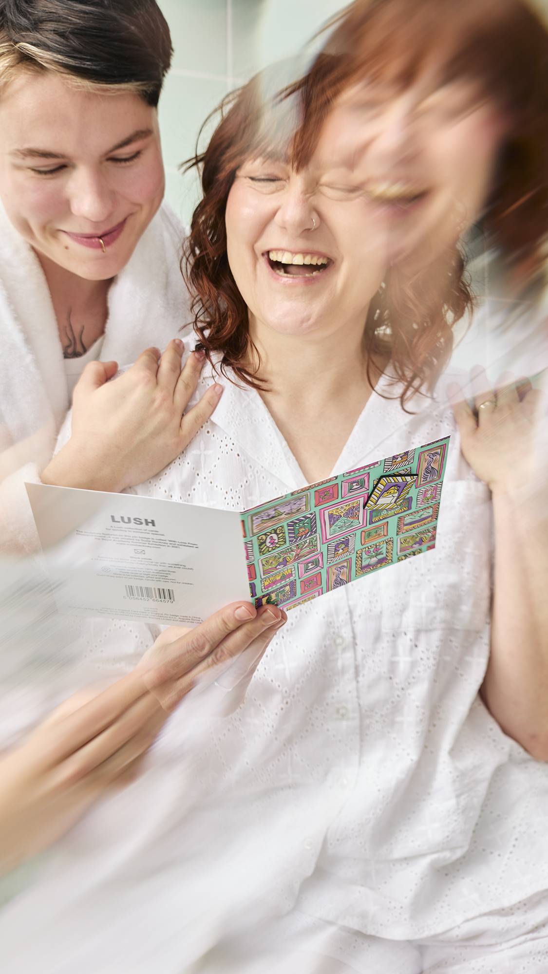 Two models in white bathrobes are laughing as they hold open a "Pictures For You" greetings card. The card features a colourful, hand-drawn gallery wall pattern. One framed image on the card contains a small, removable pin badge.