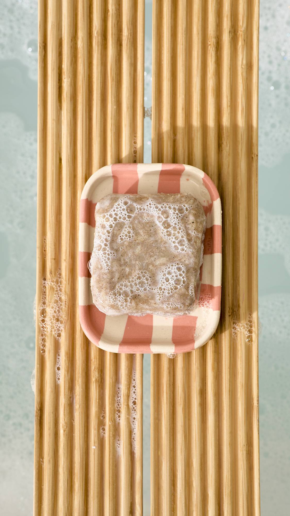 A top-down view of a bar of soapy, textured bar soap in a pink and cream chequered ceramic dish, resting on a slatted wooden bath caddy over foamy bathwater.