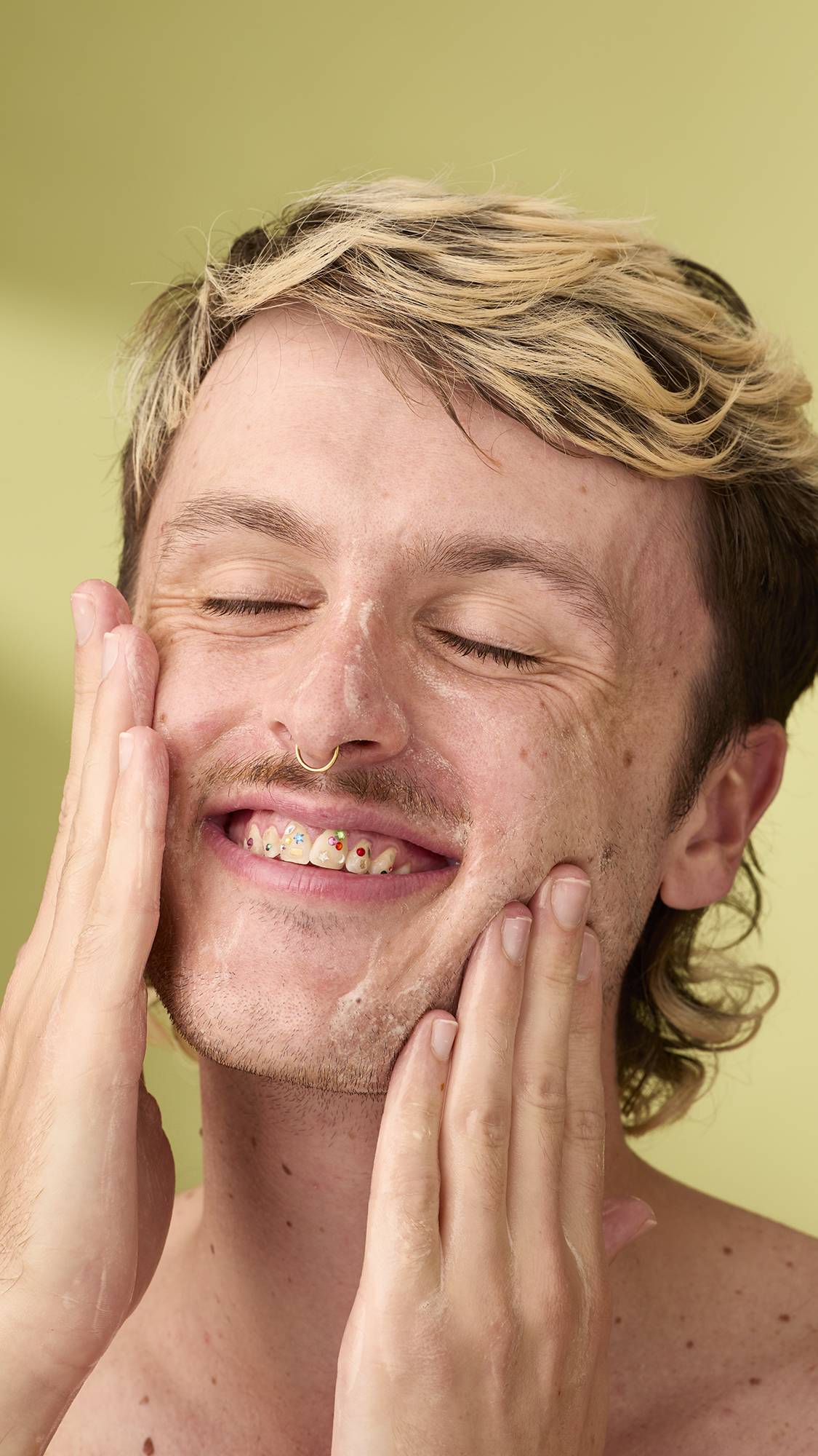A close-up of a grinning person with blonde-highlighted hair and eyes closed, washing their face with white foam from a pumpkin facial gummy. Their teeth feature colourful gem decorations against a light olive-green background.