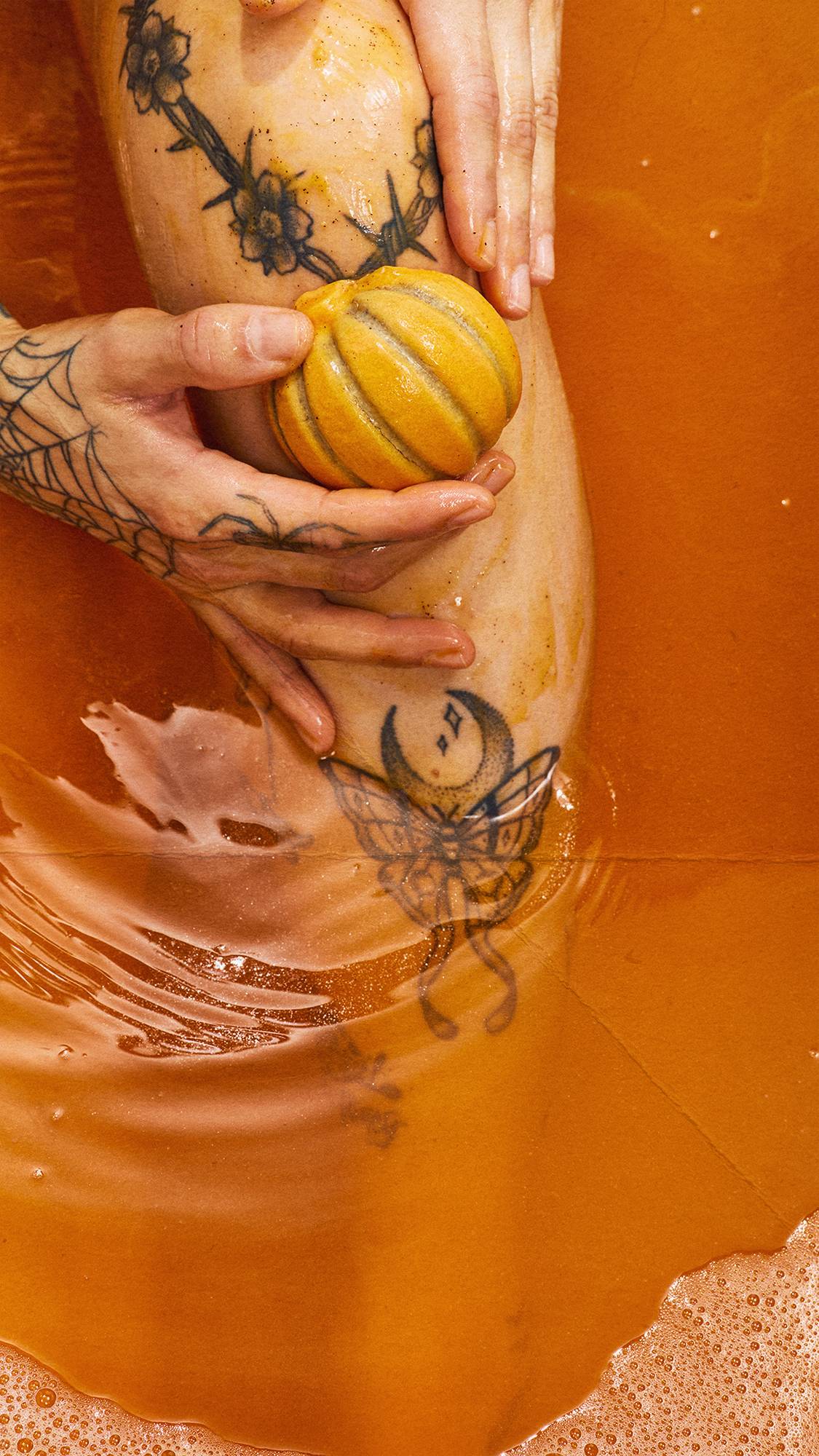 Tattooed hands smooth an orange, pumpkin-shaped solid body scrub over a leg submerged in warm, amber-coloured bathwater filled with soft bubbles.