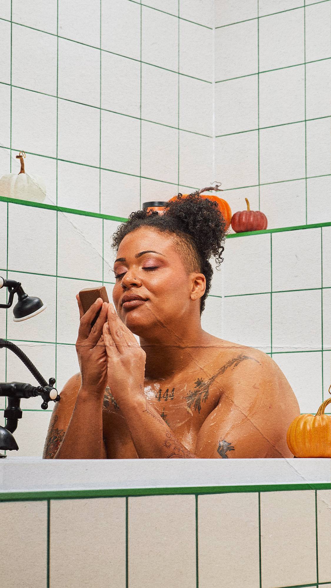 A tattooed person sits in the tub with eyes closed, gently holding an orange bar of soap to their nose. The white-tiled bathroom with green grout is decorated with colourful pumpkins.