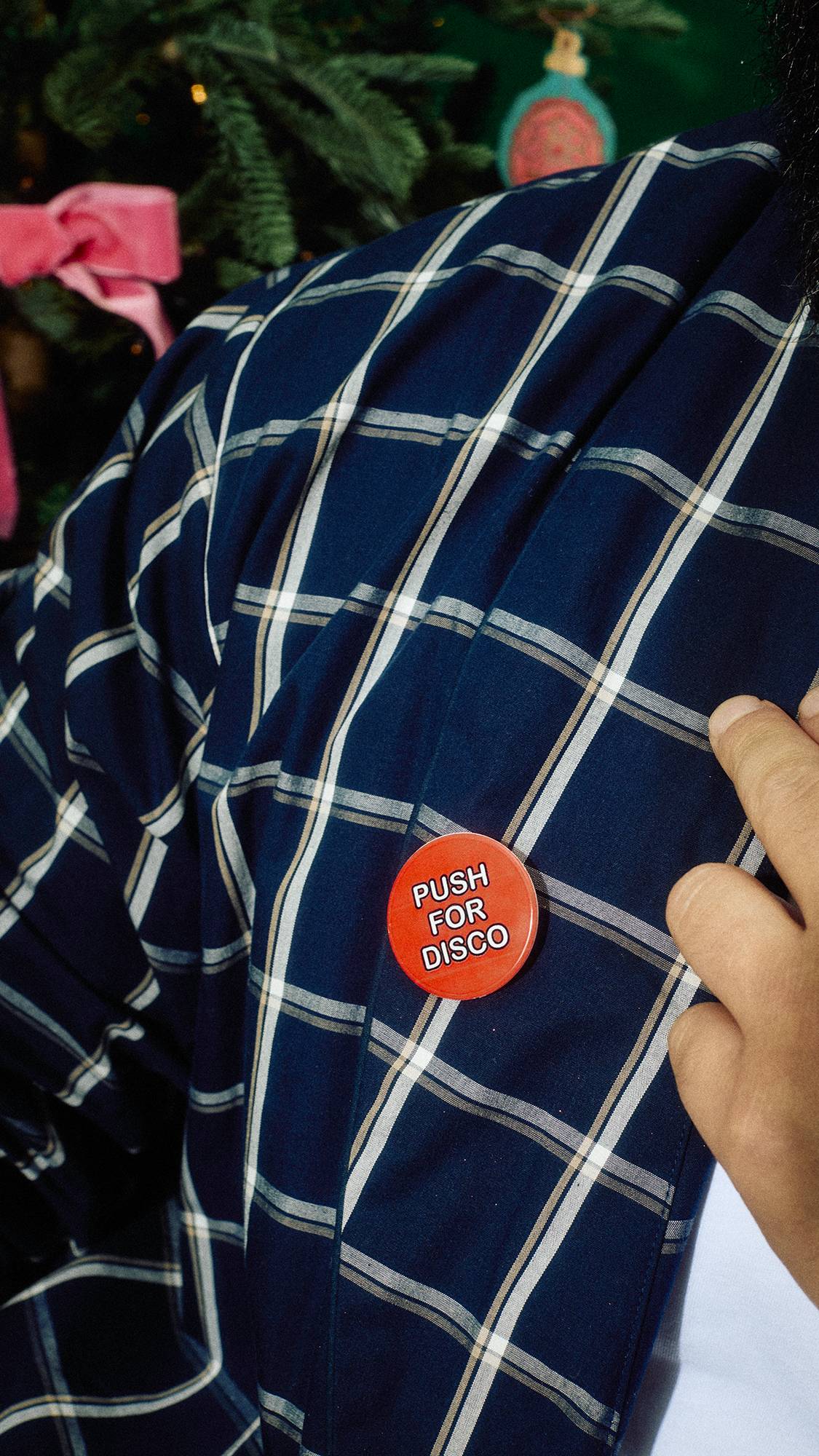 A close-up of a person wearing navy blue plaid pyjamas, with a hand touching a round red badge pinned to the chest that reads "Push for Disco" in white text, set in front of a Christmas tree.
