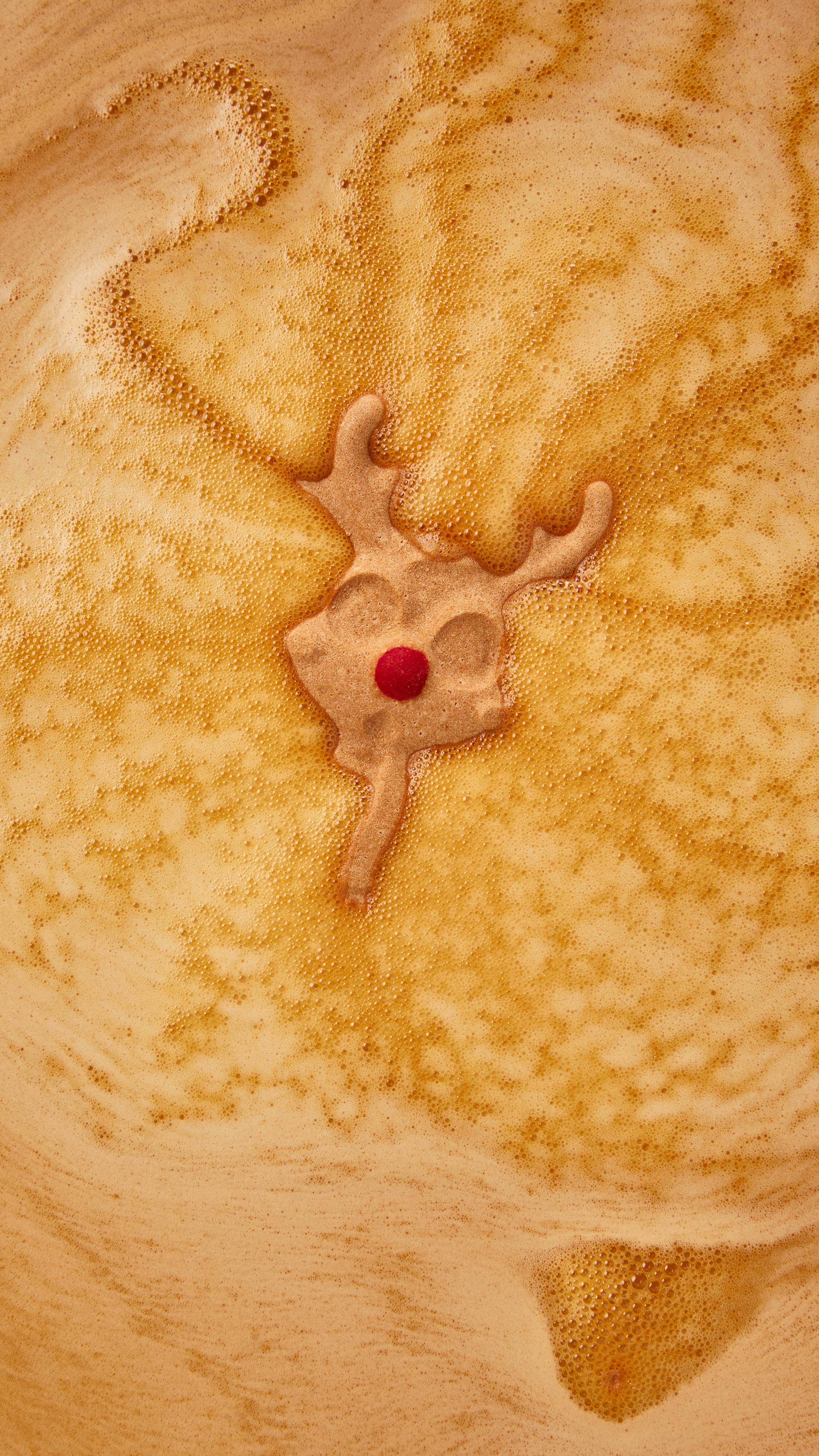 A partially dissolved reindeer bath bomb with a prominent red nose floats amidst swirling patterns of golden, creamy bath foam.