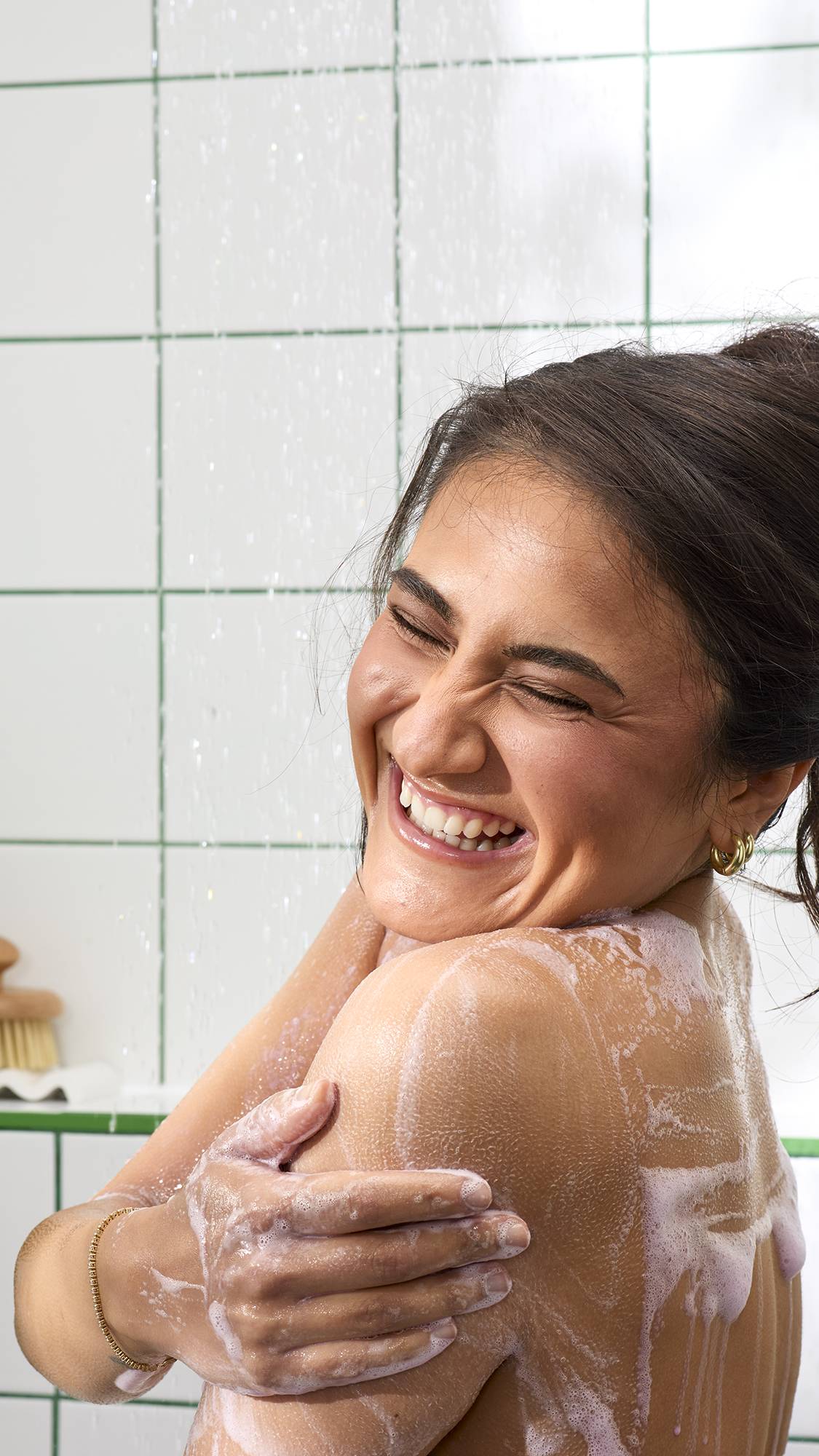 A close-up shot of a laughing person with their hair tied up, washing their shoulder in the shower. A light pinkish, frothy soap lather coats their skin against a background of white tiles with green grouting.