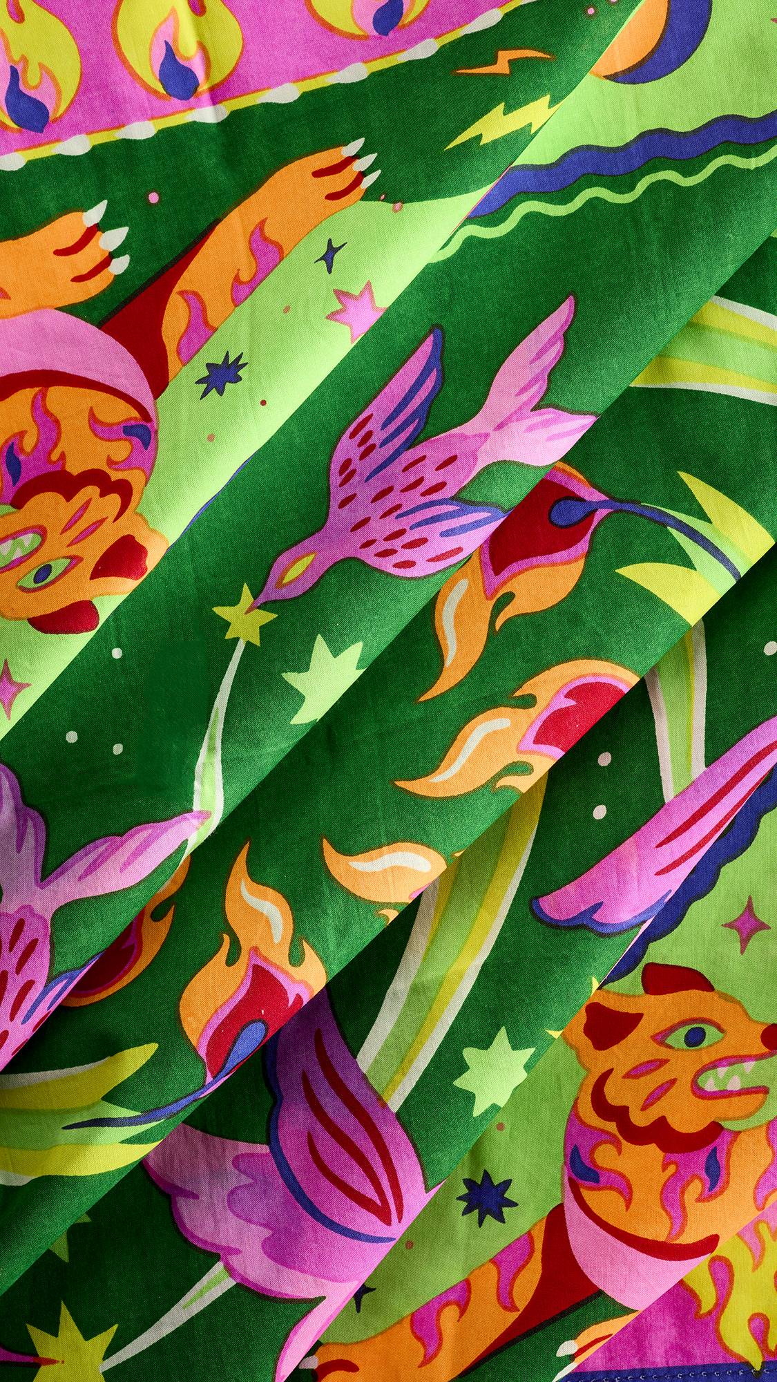 A close-up shot of folded green fabric from a Lush "Roar and Shine" Knot Wrap, featuring intricate printed illustrations of pink birds, orange tigers, flames, and stars.