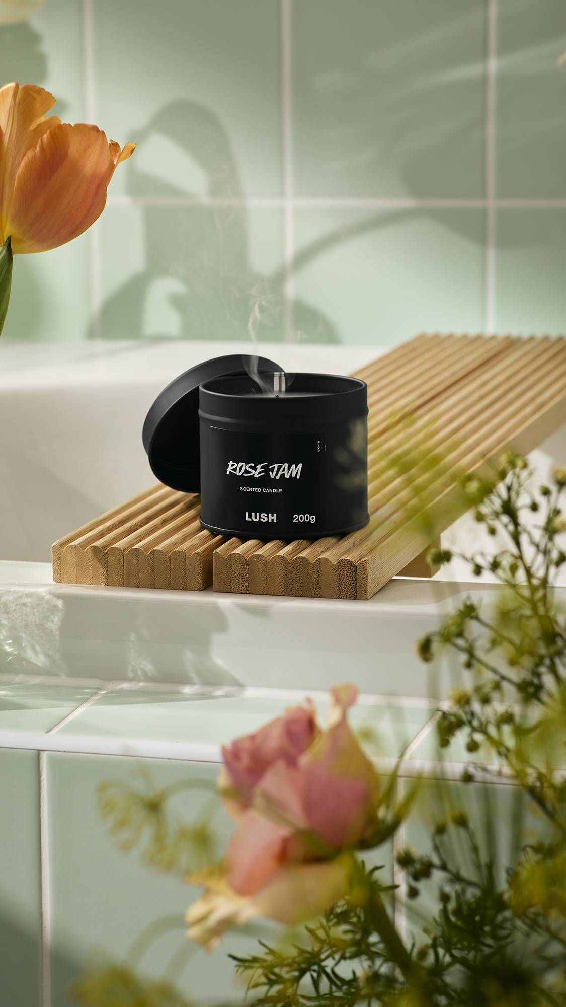 A close-up shot shows a Lush "Rose Jam" scented candle in a round, black tin with its lid leaning against it. The candle has been extinguished, and a thin wisp of white smoke curls upwards. It sits on a wooden bath bridge amidst fresh flowers.