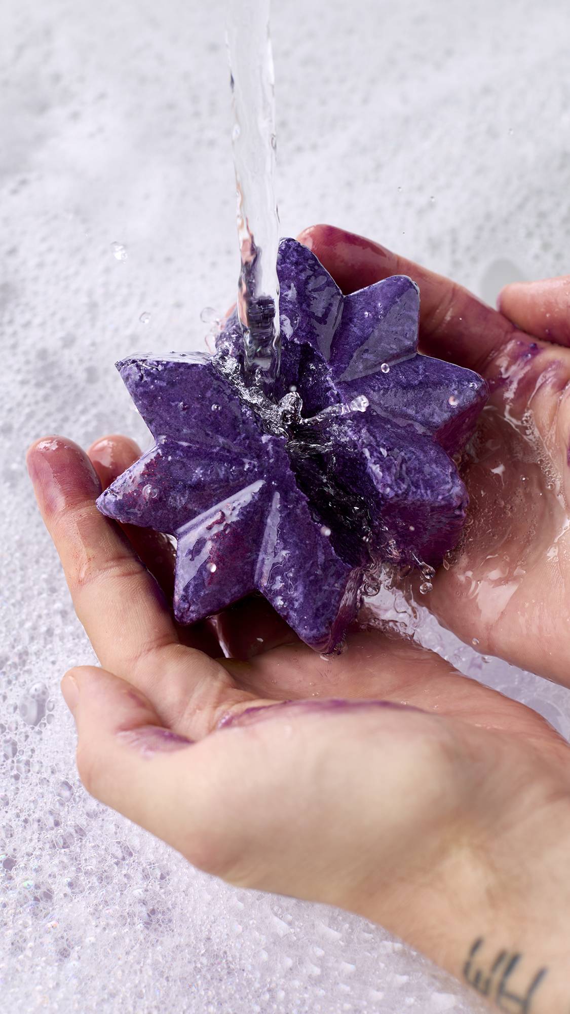 Two hands crumble a deep purple star-shaped bubble bar under running tap water over bathwater covered in dense white bubbles, releasing purple colour and shimmering lather.