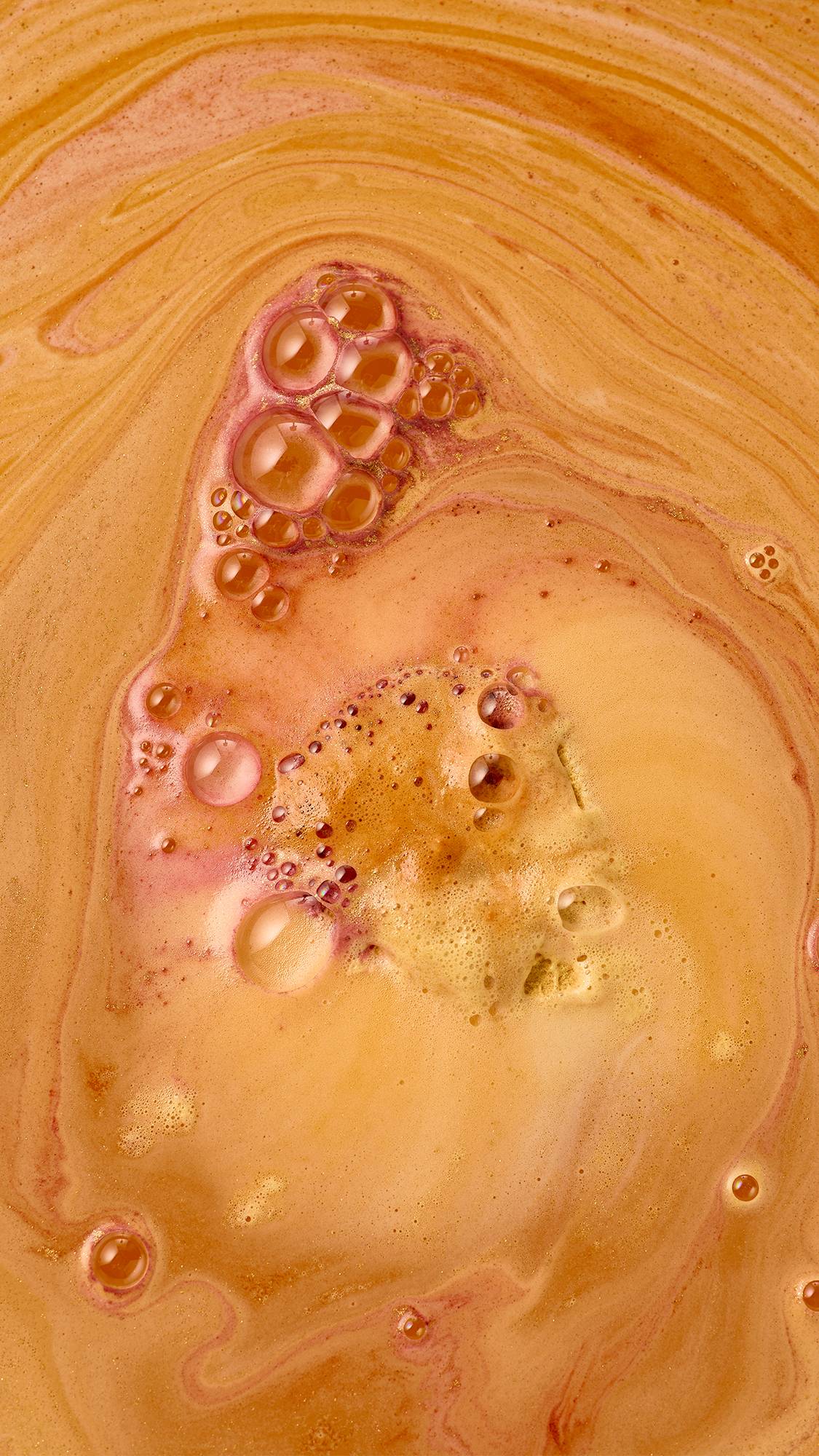 A close-up view of warm orange bathwater swirling with golden shimmer, creating bubbly foam and delicate pink ribbons of colour as a Lush bath bomb dissolves.