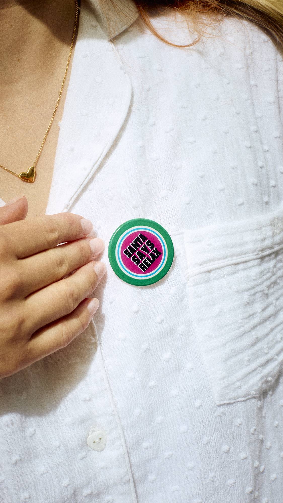 A close-up of a person wearing white pyjamas, touching a round green badge pinned to the chest that reads "Santa Says Relax" in black text on a pink centre.