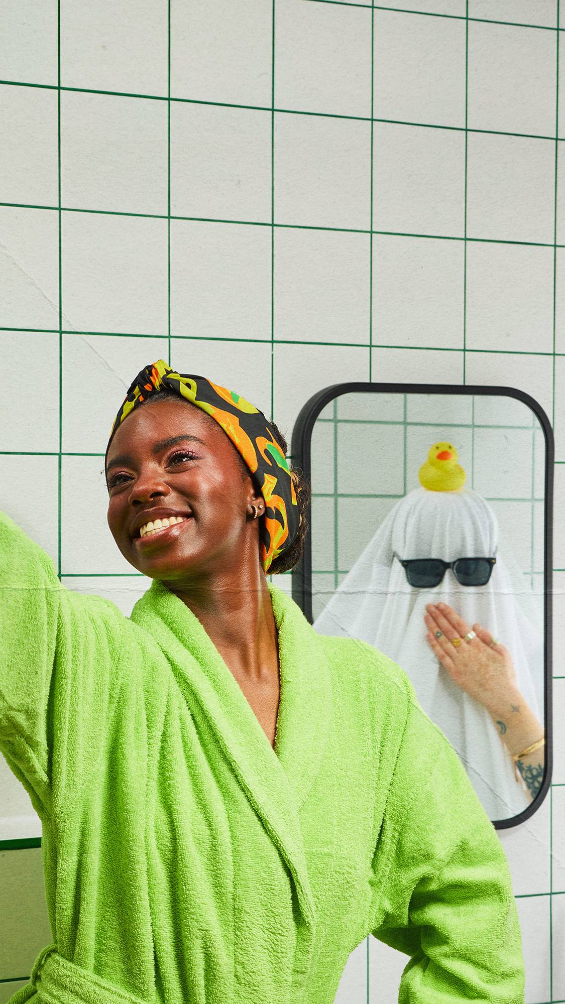 A model in a lime green dressing gown and patterned headscarf, fashioned from a knot wrap, takes a selfie in a bathroom. Their reflection in the mirror shows a person dressed as a ghost under a white sheet, wearing sunglasses with a rubber duck.