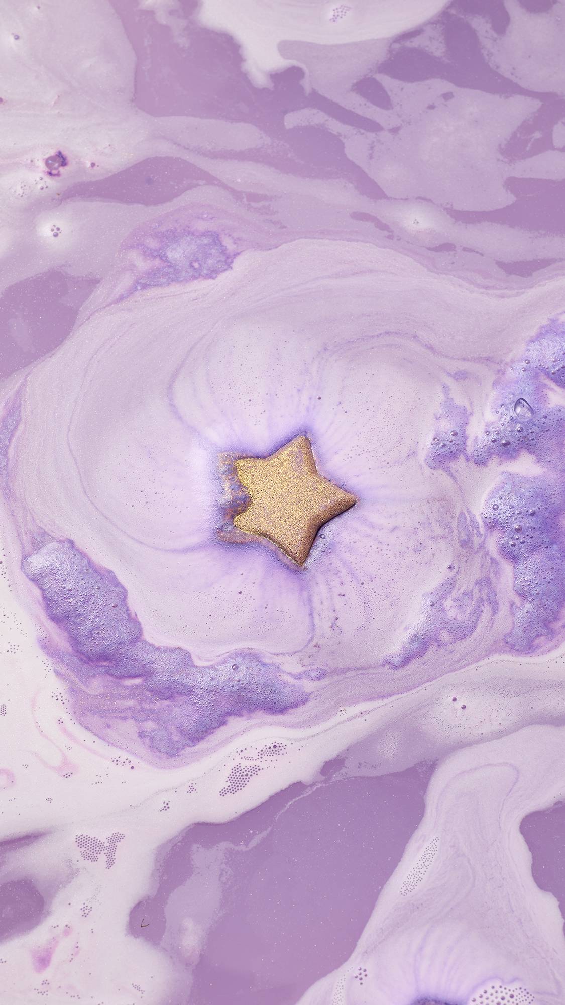  The gold-glittered, star-shaped Siraj bath bomb fizzes and dissolves in the center of the bath, creating vibrant swirls of purple water and thick lilac and white foam on the surface.