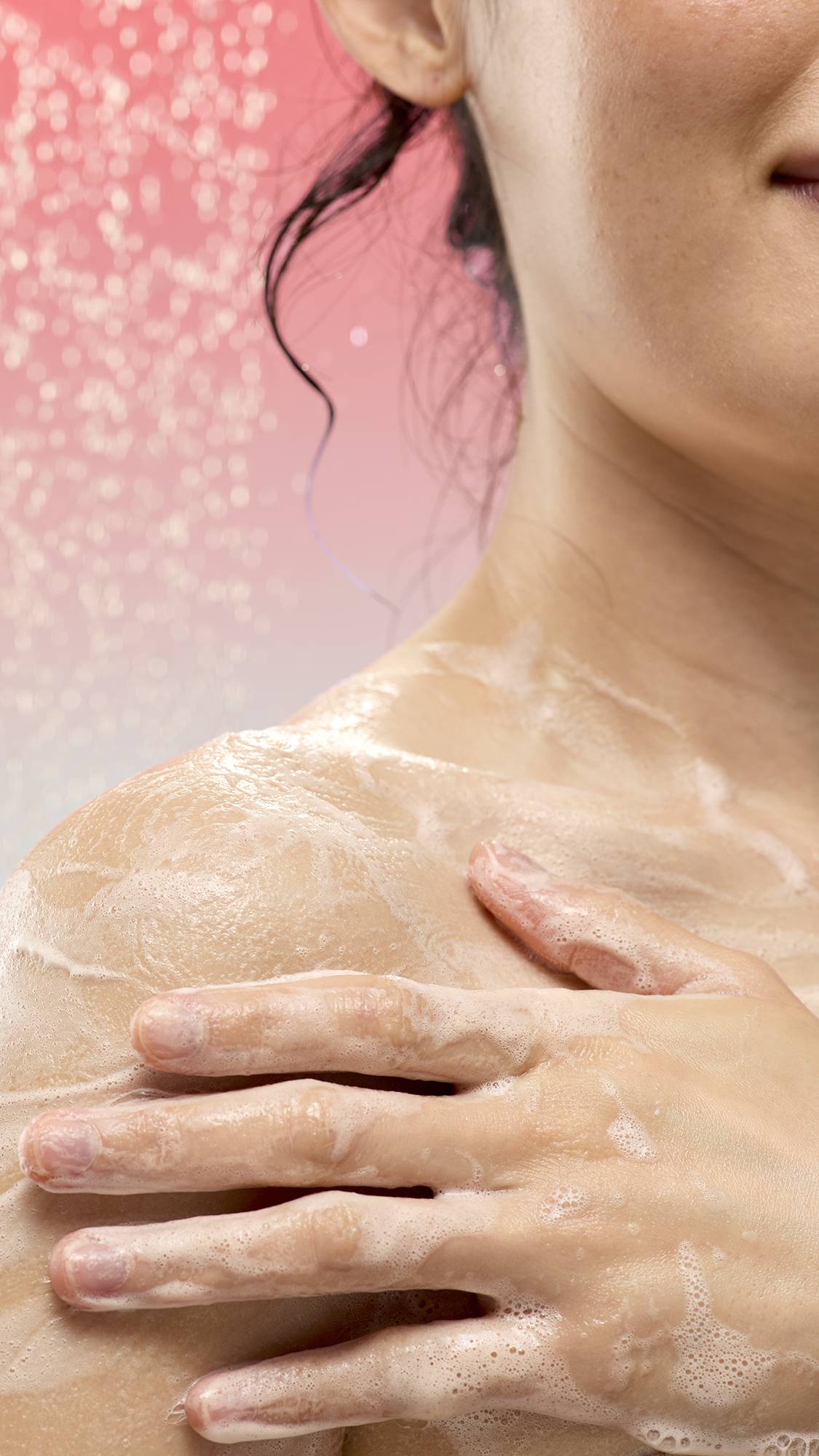 A close-up of a person applying foamy white lather from Smell Of Roses soap to their shoulder. Falling water droplets spray in the background against a soft pink-to-blue gradient.