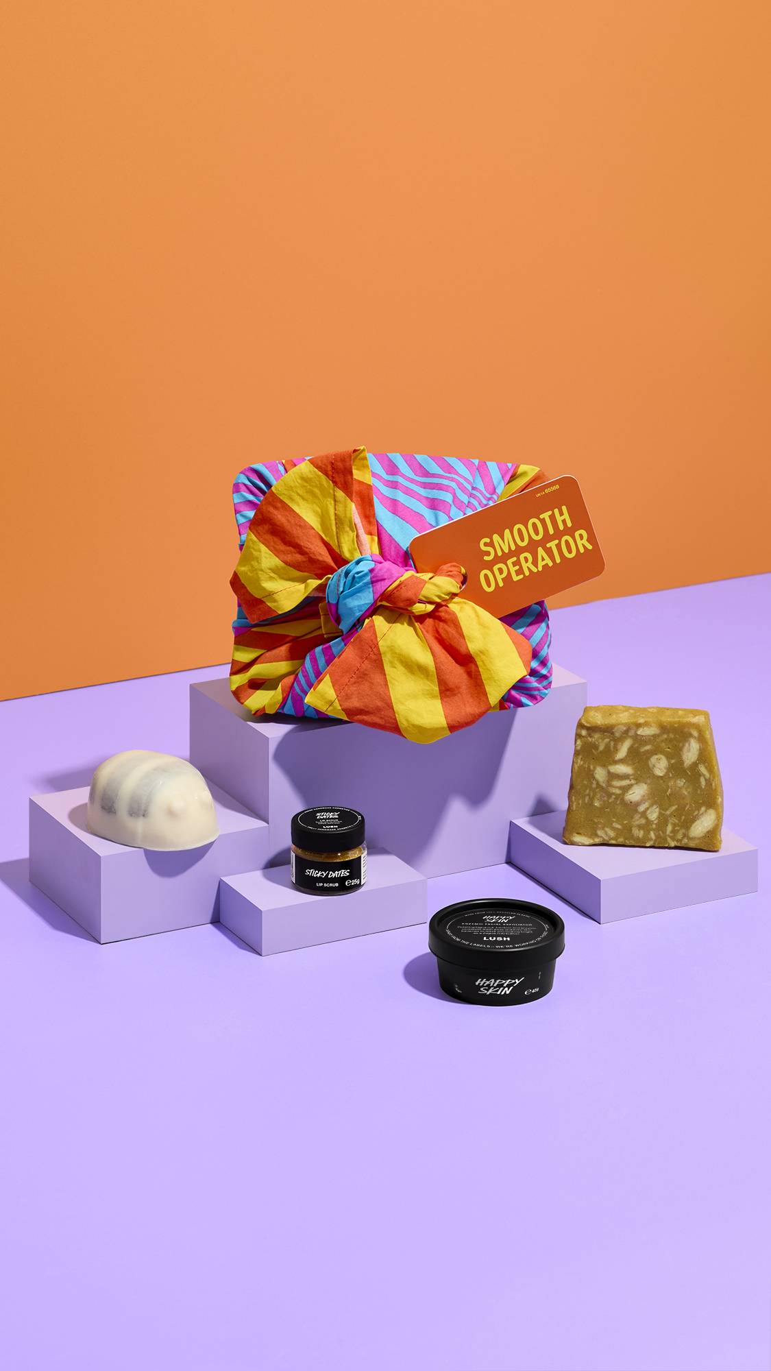 A brightly patterned knot-wrapped gift tagged "SMOOTH OPERATOR" sits atop lilac plinths against an orange background, surrounded by a scrubee solid body scrub bar, Sticky Dates lip scrub, Happy Skin facial exfoliator, and a bar of soap.