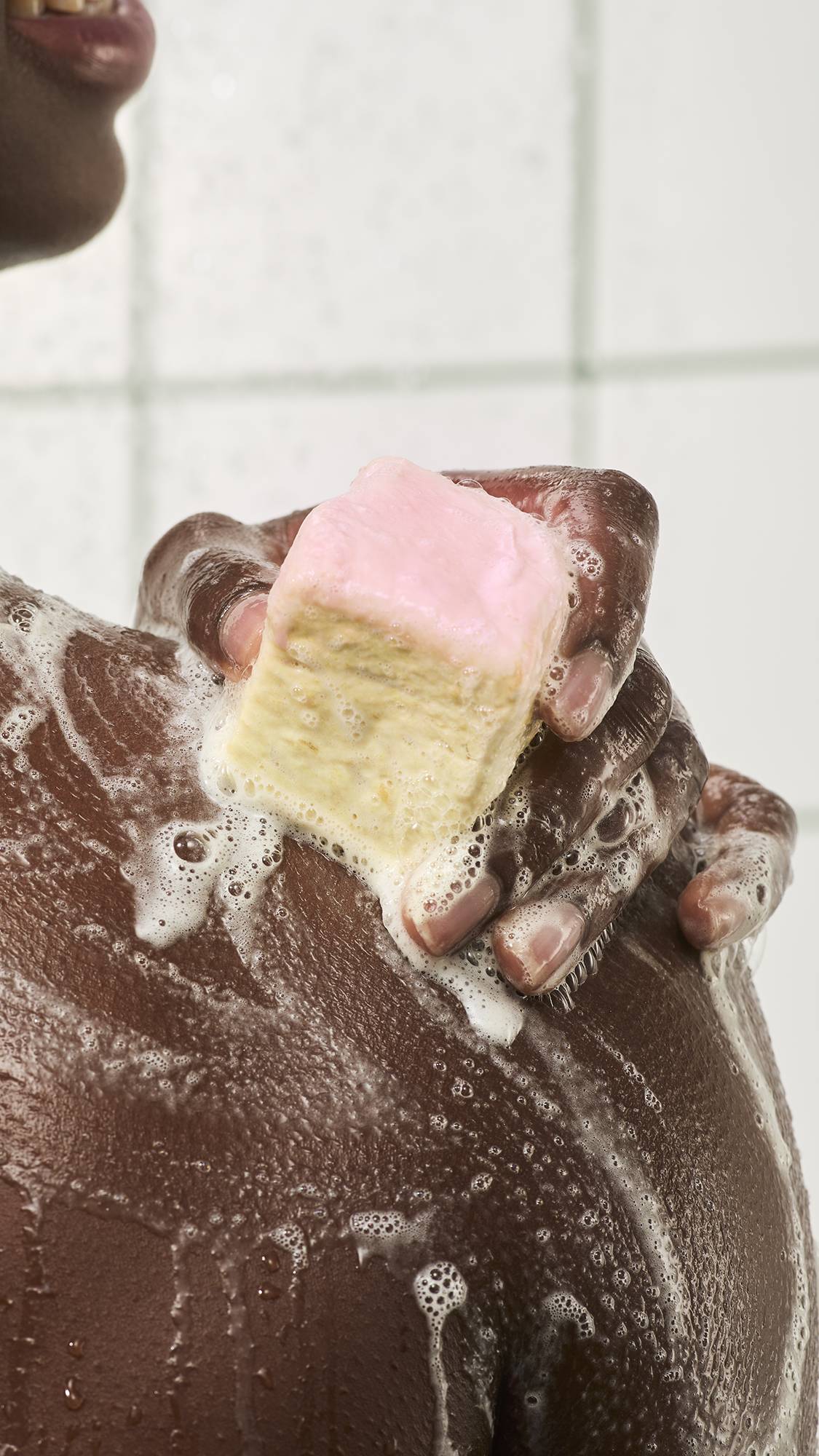 A person lathers a square "S'mores" soap with a smooth pink top layer over their shoulder in the shower, spreading white bubbly suds across wet skin against a white tiled background.
