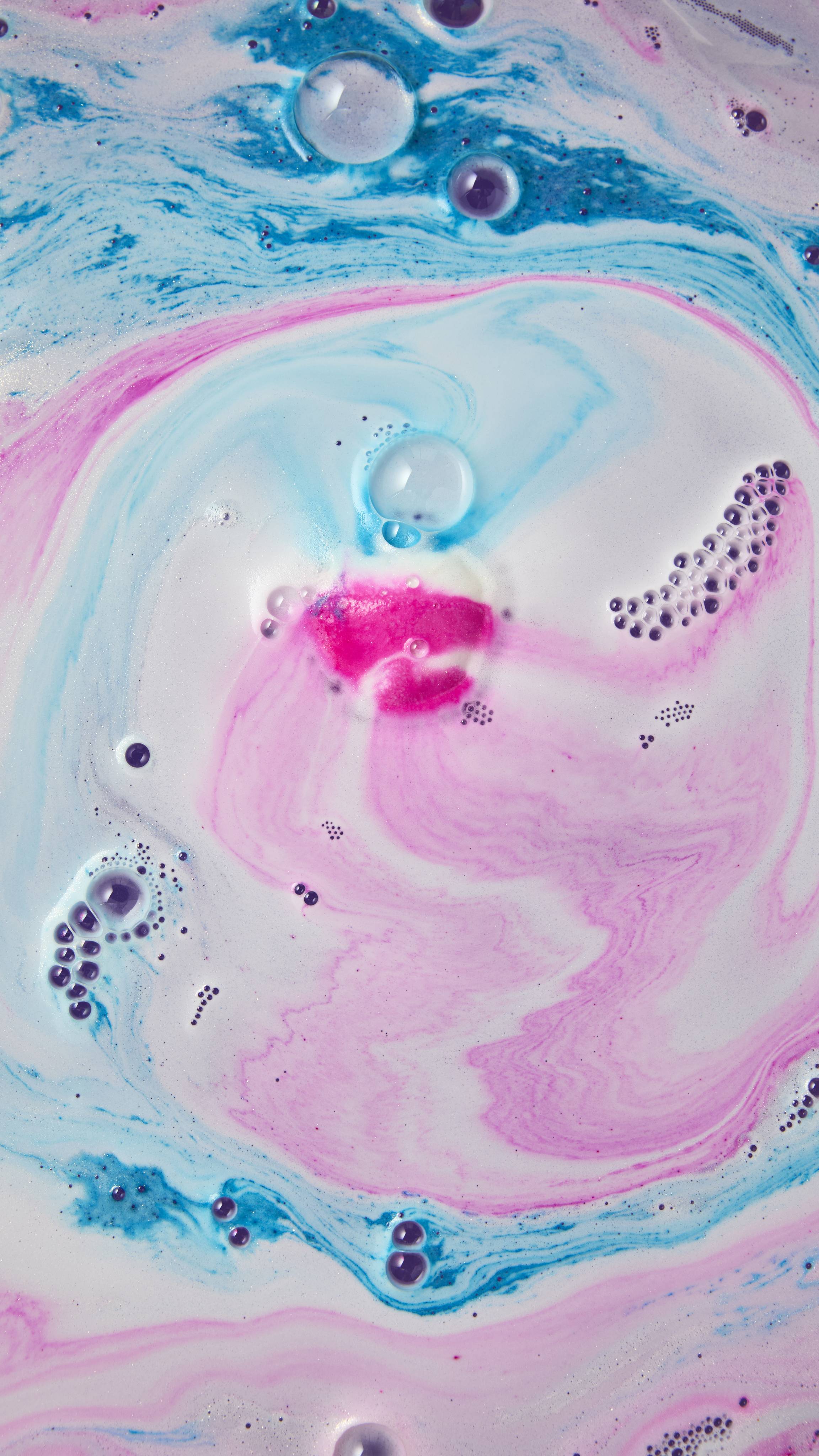 A "Snow Fairy" bath bomb dissolves into a pool of soft white and pastel pink foam, surrounded by vibrant swirls of blue and magenta bathwater with scattered bubbles.