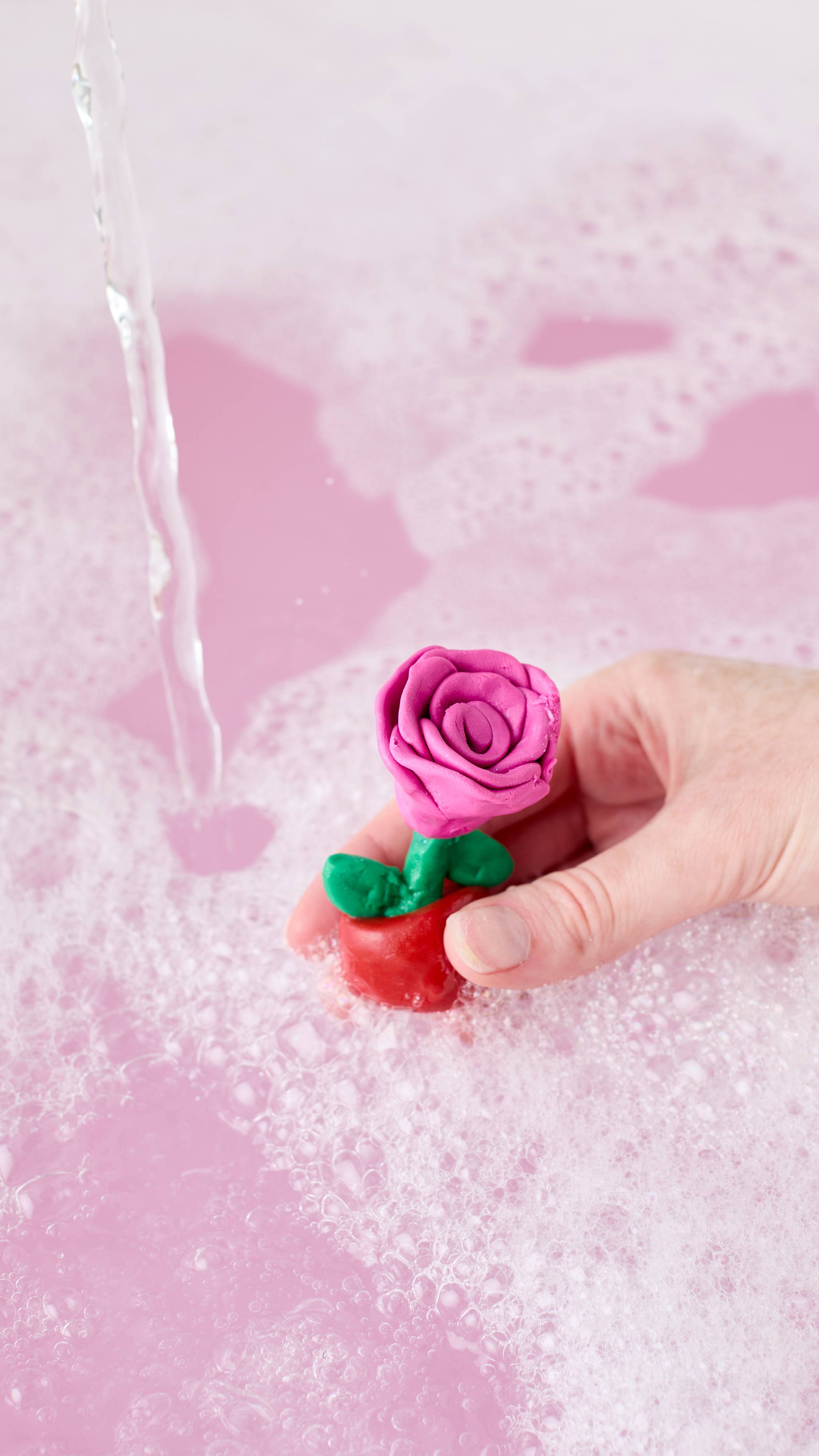 A hand holds a small rose-shaped model crafted with the "Snow Fairy" FUN. It has vibrant pink petals and green leaves near running tap water, above pastel pink bathwater covered in soft white bubbles.