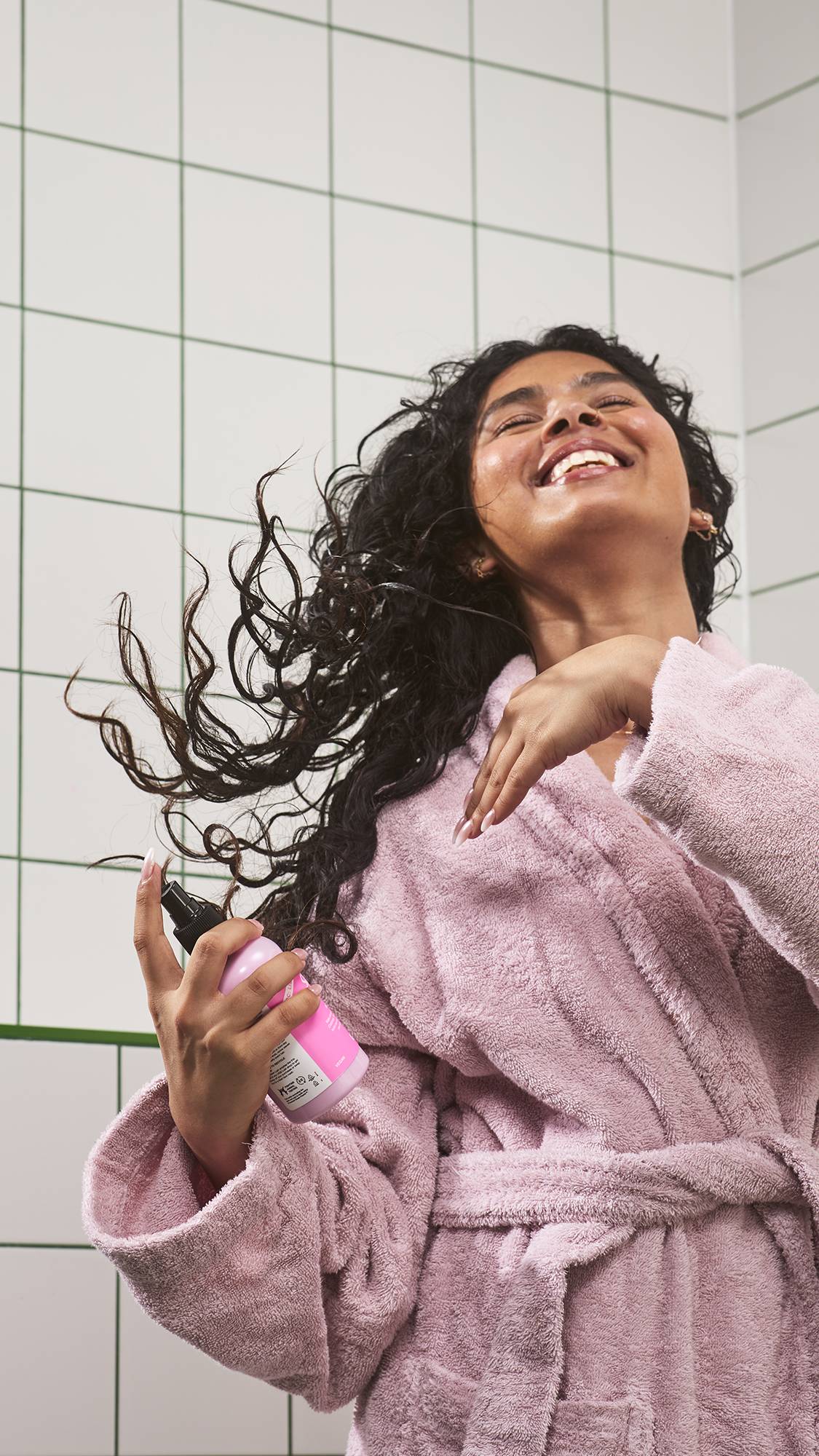 A smiling model wearing a plush pink dressing gown flips their long, dark, curly hair back while holding a bottle of Lush "Snow Fairy" spray in a white tiled bathroom.