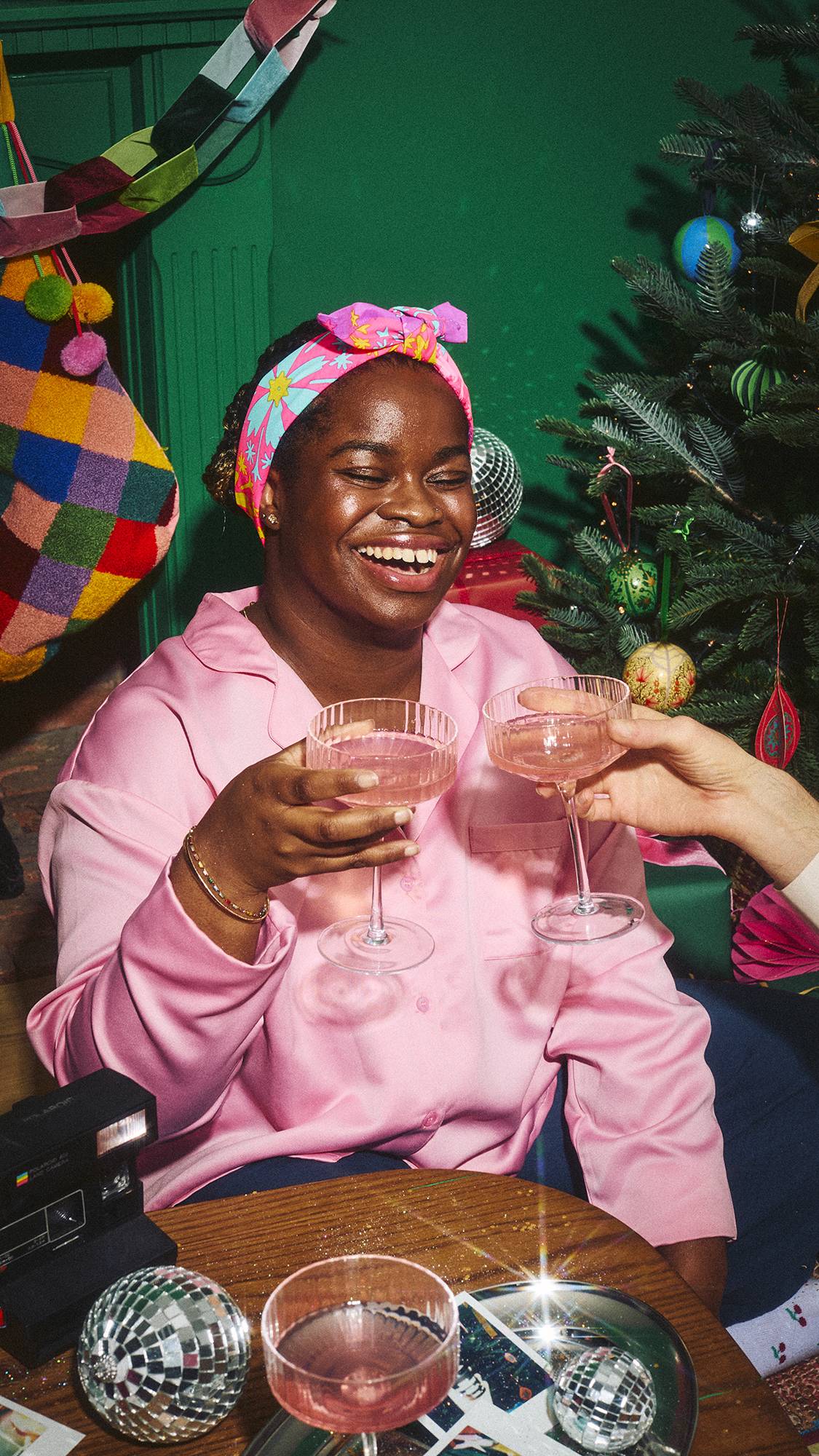 A laughing person wearing pink pyjamas and a pink floral Lush fabric Knot Wrap fashioned into a headband toasts pink drinks with a friend in a festive setting, beside a Christmas tree and a table with disco balls and cameras.