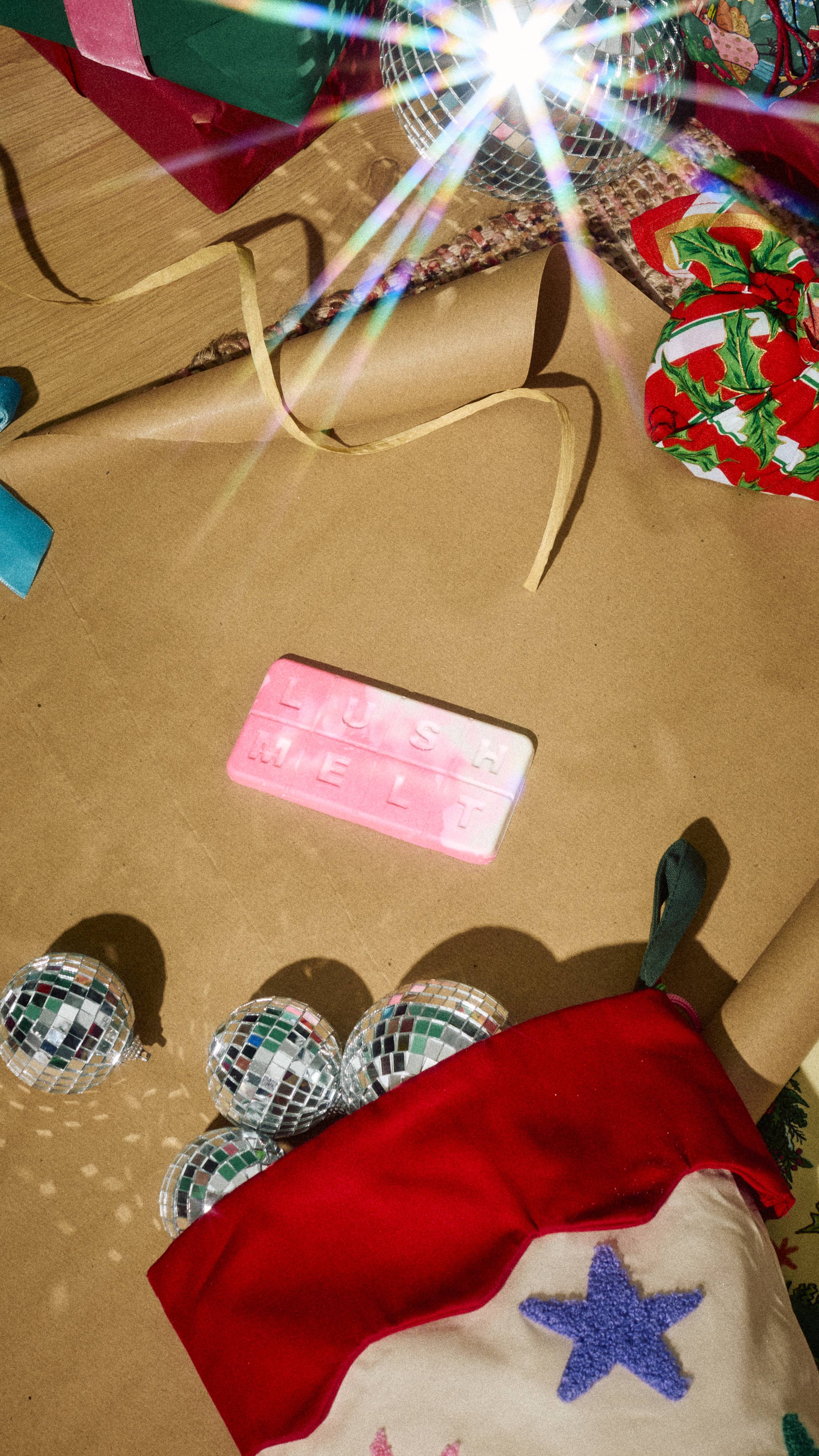 A rectangular pink and white marbled wax melt bar embossed with "LUSH MELT" lies on brown wrapping paper, surrounded by small disco ball ornaments spilling from a Christmas stocking and festive gifts.