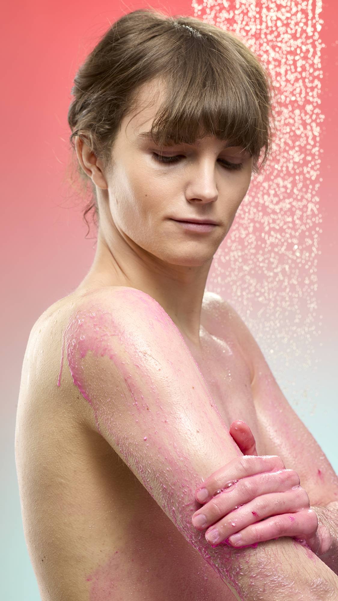 A person lathers their shoulder and arm with vibrant magenta foam from Snow Fairy Pop shower powder while standing under a spray of falling water. Set against a soft pink-to-blue gradient background.