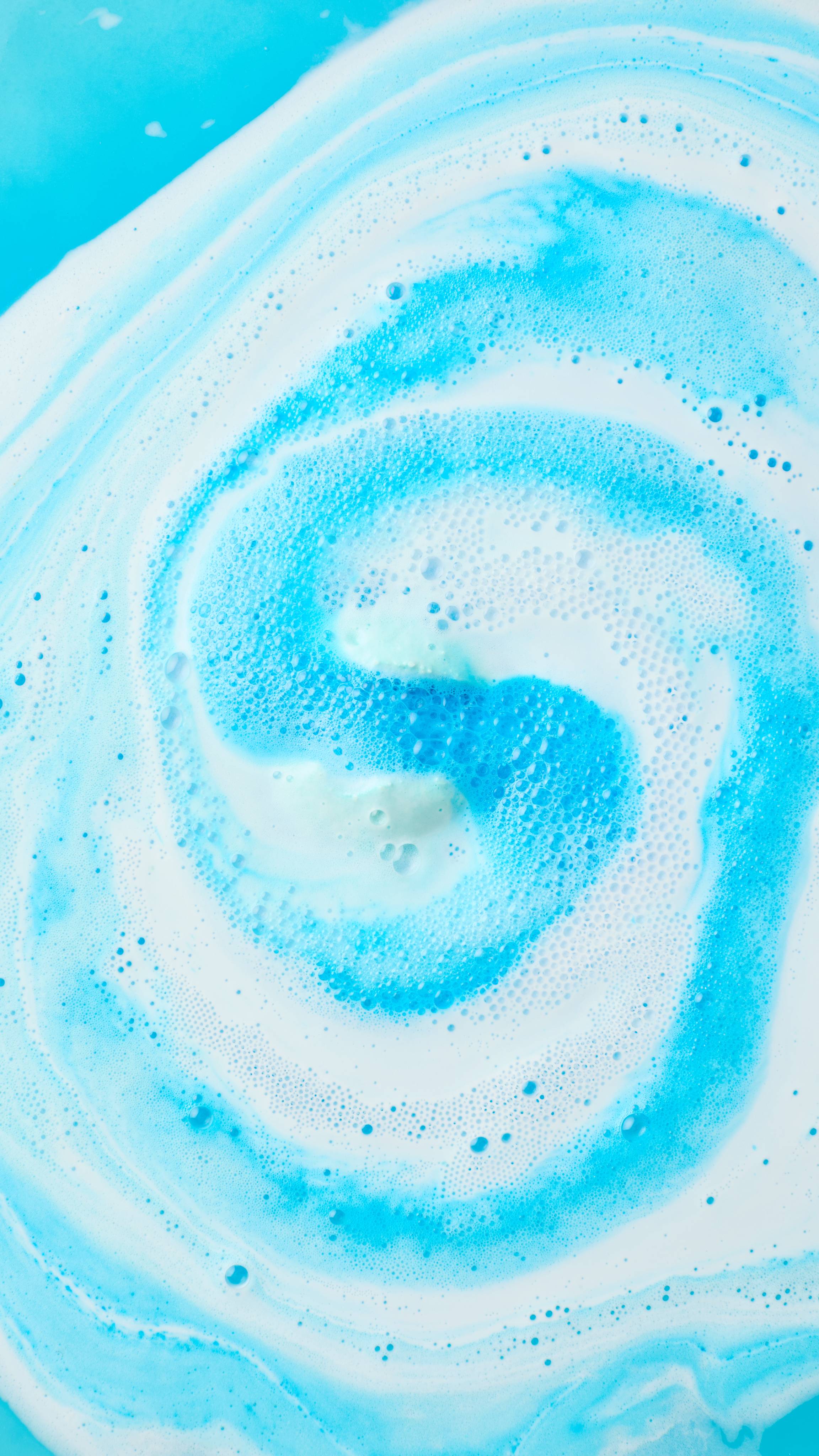 Swirling streams of bright blue and white bath foam create a dynamic, spiral pattern across vibrant cyan bathwater as a bath bomb dissolves at the centre.