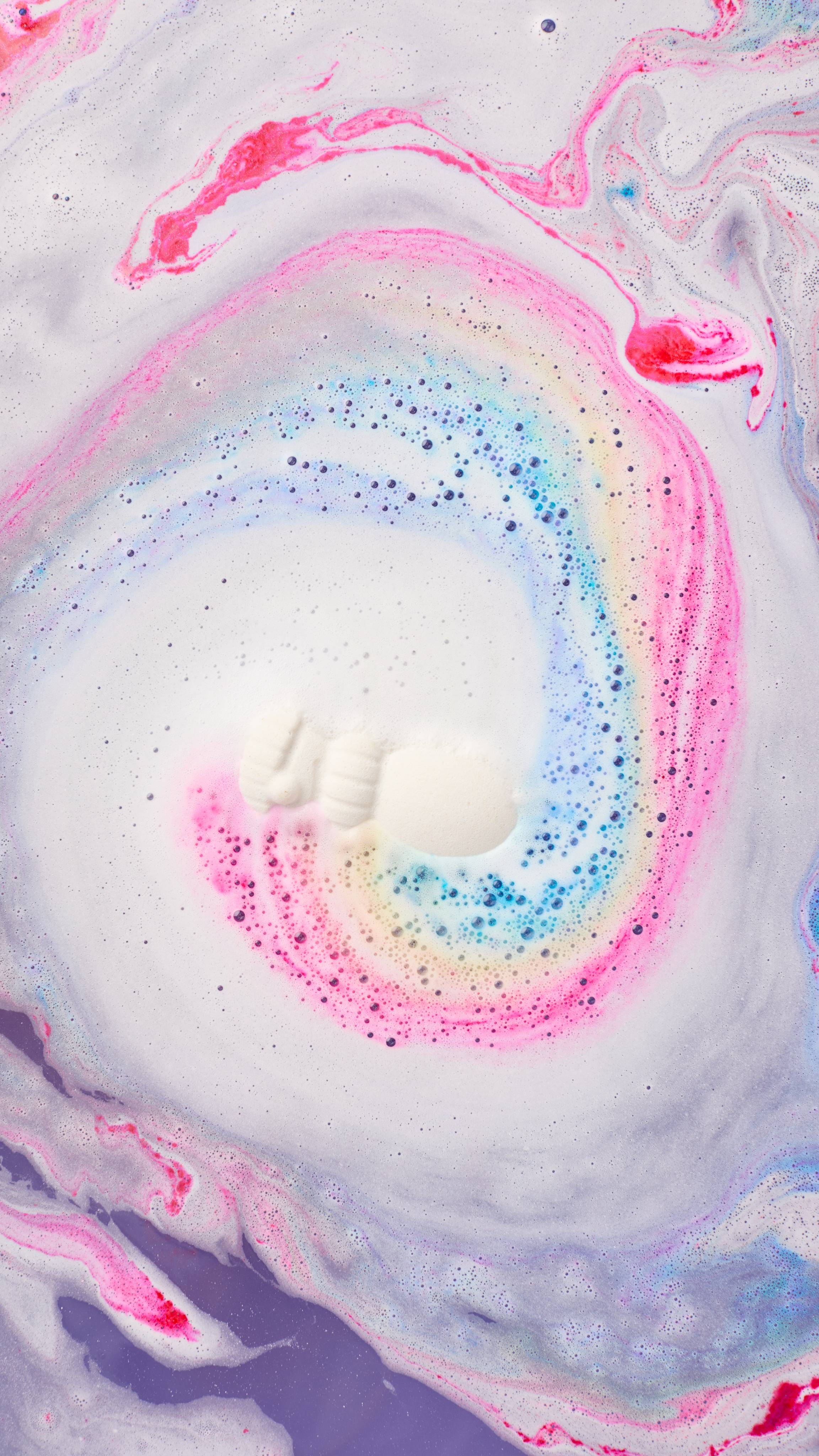 A white snowman bath bomb dissolves at the centre of bathwater, spinning into a vibrant rainbow swirl of pink, blue, yellow, and purple frothy foam.