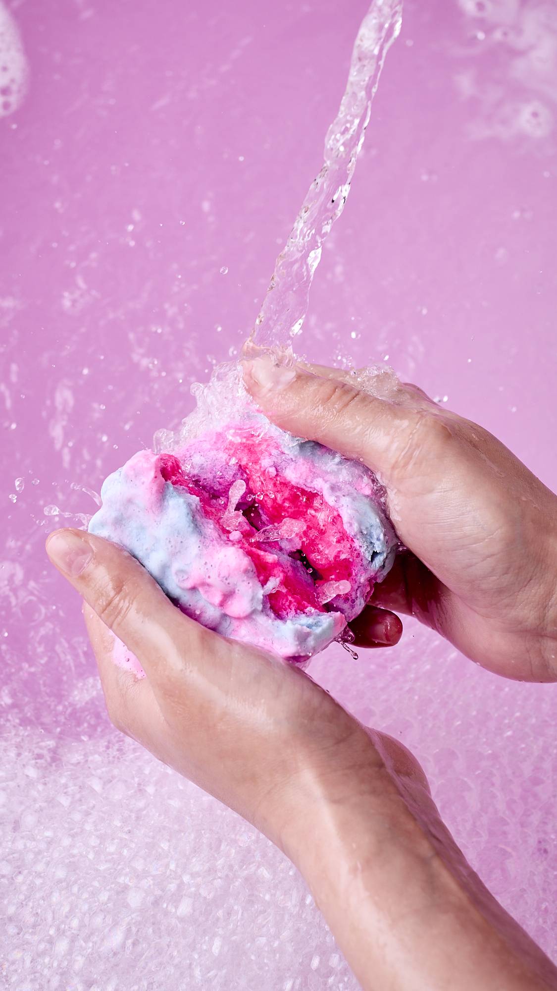A close-up shot of hands breaking apart a dissolving, mooncake-shaped bubble bar under running water. The split reveals a vibrant pink centre, splashing into a bath filled with frothy pink bubbles.
