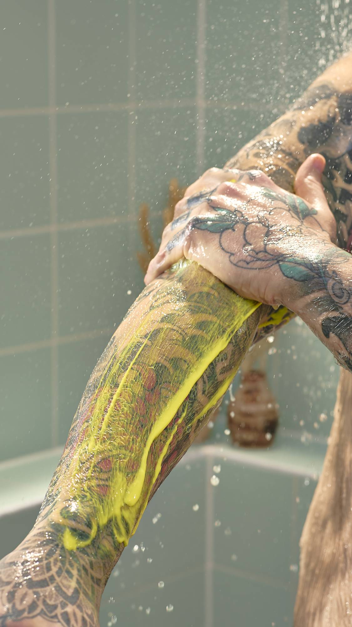 The image shows a close-up of a model's tattooed arm in a shower, with bright yellow "Squeeze The Day" shower gel being spread across the skin. Water droplets fall in the background against pale green wall tiles.