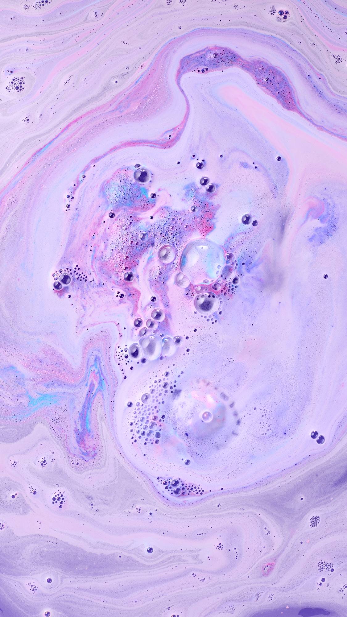 A bath bomb dissolves into a pool of soft lilac and lavender bathwater, creating vibrant patches of purple, magenta, and periwinkle blue fizz with clusters of glossy, clear bubbles.