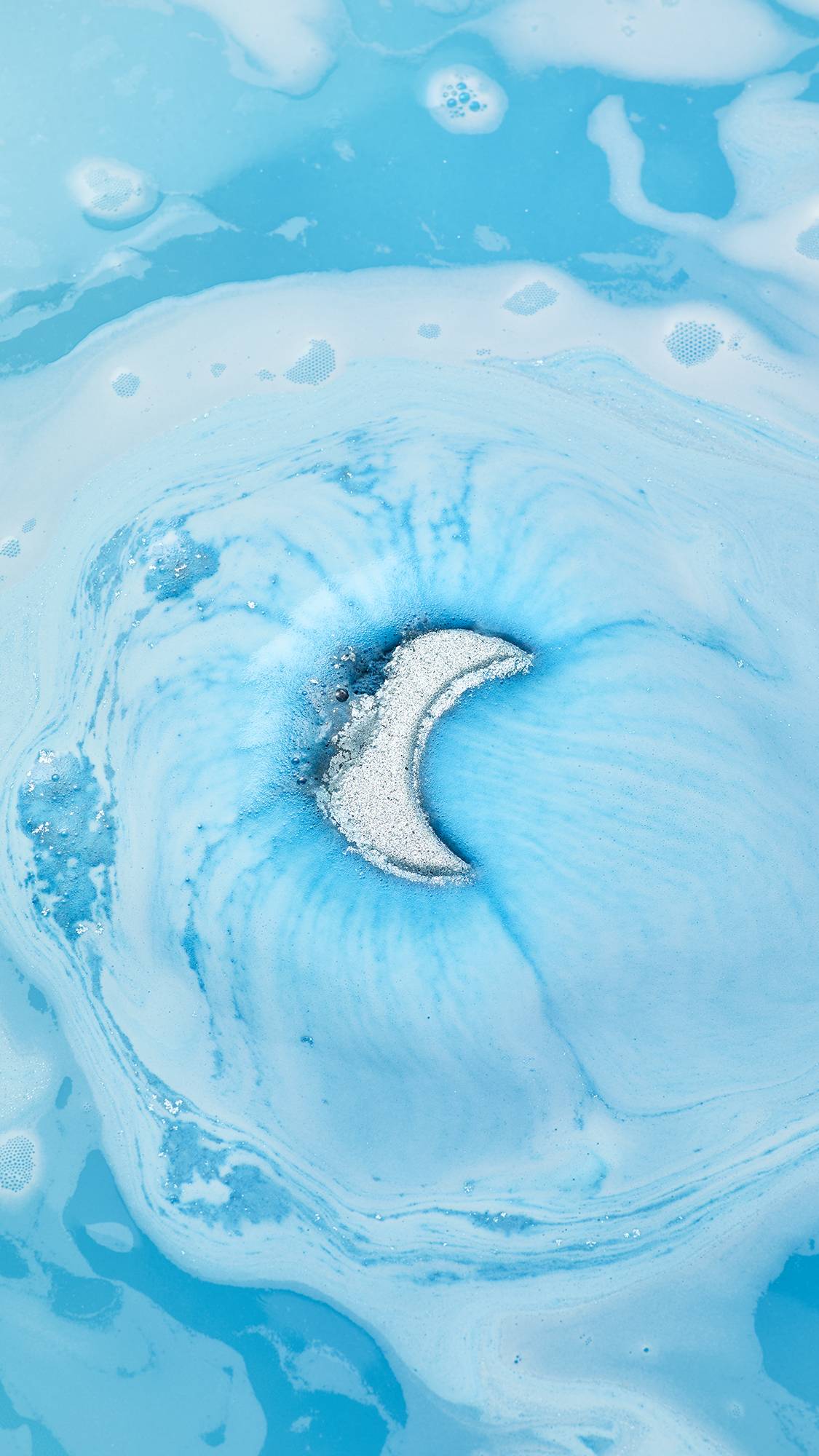 The shimmering silver-blue Sukoon bath bomb, shaped like a crescent moon, fizzes and dissolves in bright blue water, creating vibrant swirls of light blue and white foam on the surface.