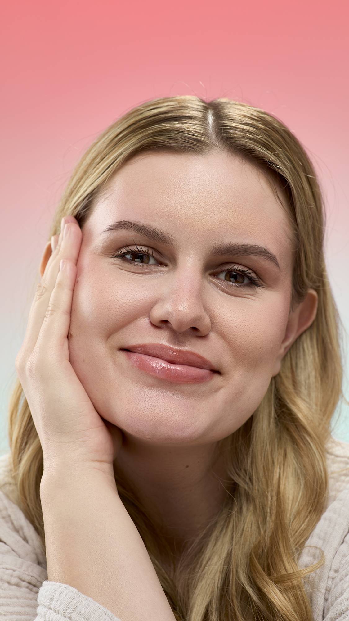 A smiling person with blonde wavy hair rests their face on their hand, showing a glossy finish on their lips from Super Milk lip gloss. They are wearing a light, textured robe against a pink gradient background.