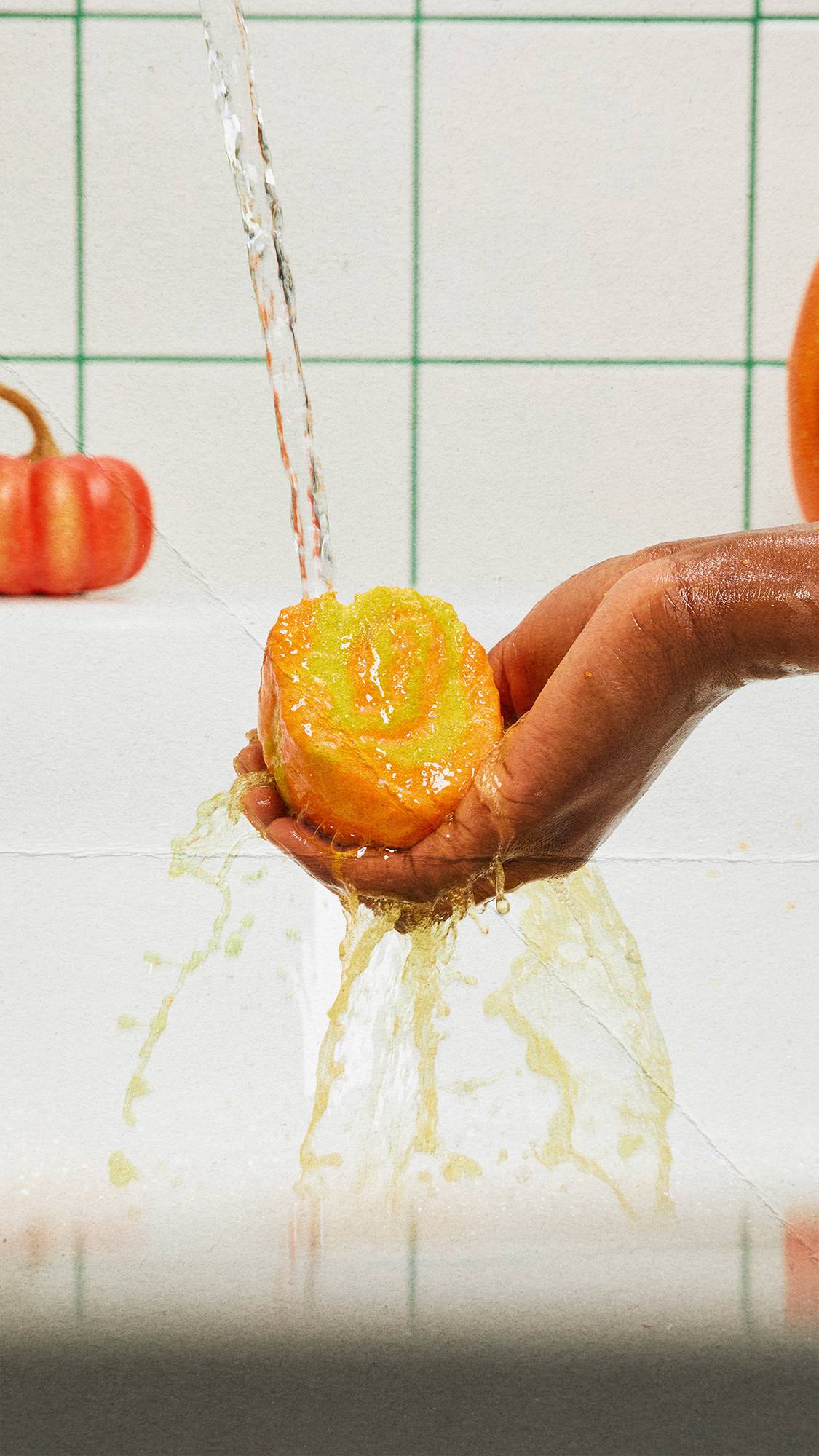 A hand holds an orange and yellow swirled bubble bar directly under running tap water, with yellow water splashing down against white-tiled bathroom walls featuring green grout and a small orange pumpkin in the background.