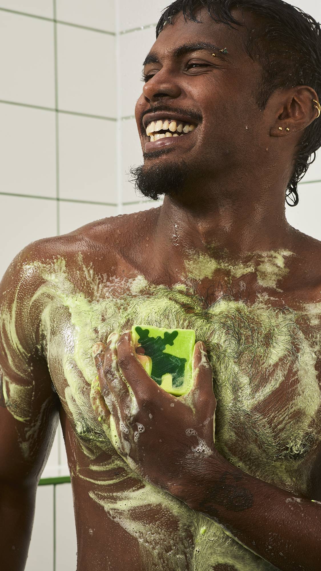 A smiling model lathers a square lime green soap featuring a green reindeer design across their chest, spreading soft greenish-white foam on wet skin in the shower against a white tiled wall.