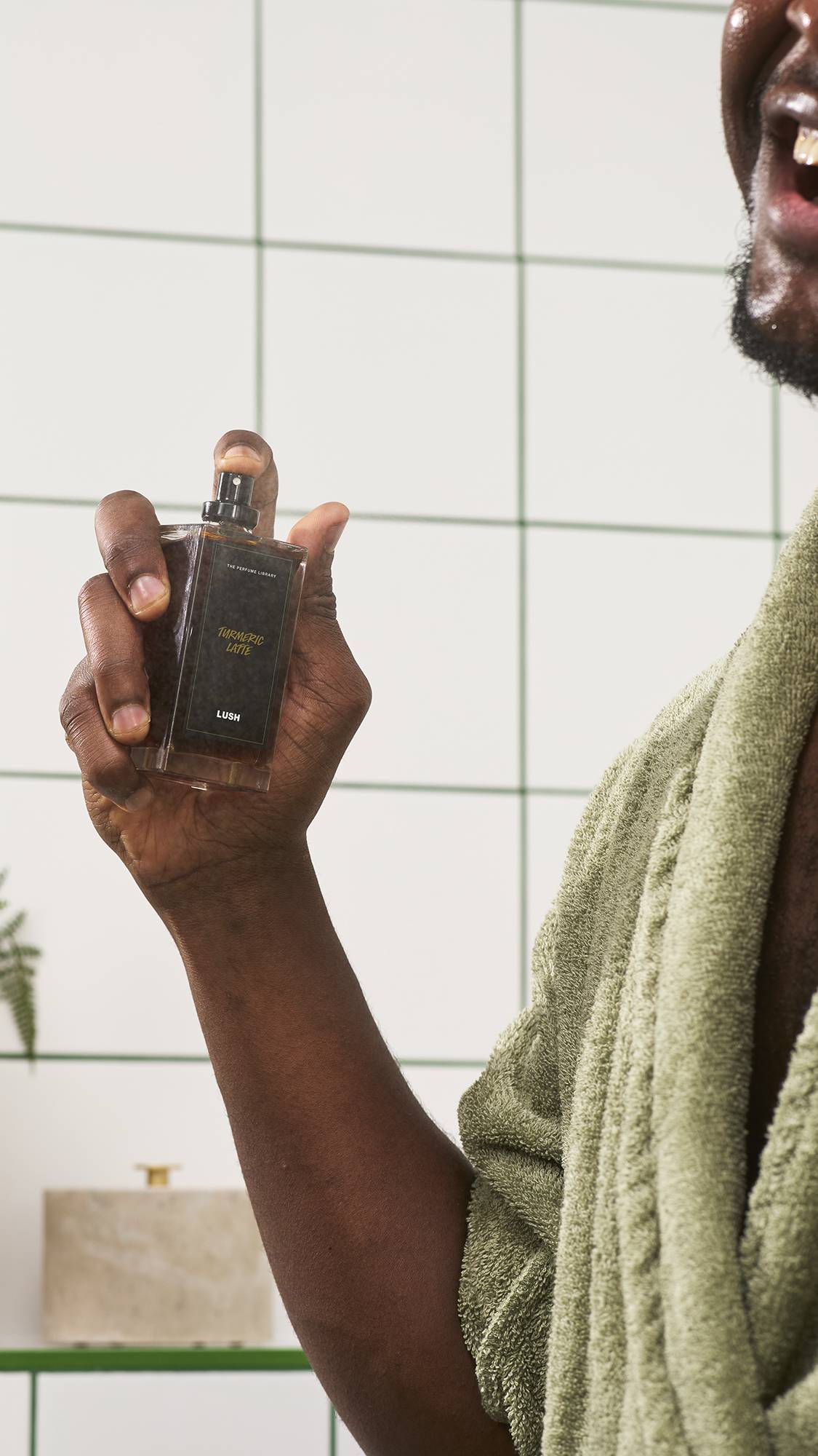 A smiling model in a green dressing gown holds up a glass bottle of Lush Turmeric Latte perfume, spraying it into the air against a white tiled bathroom wall background.