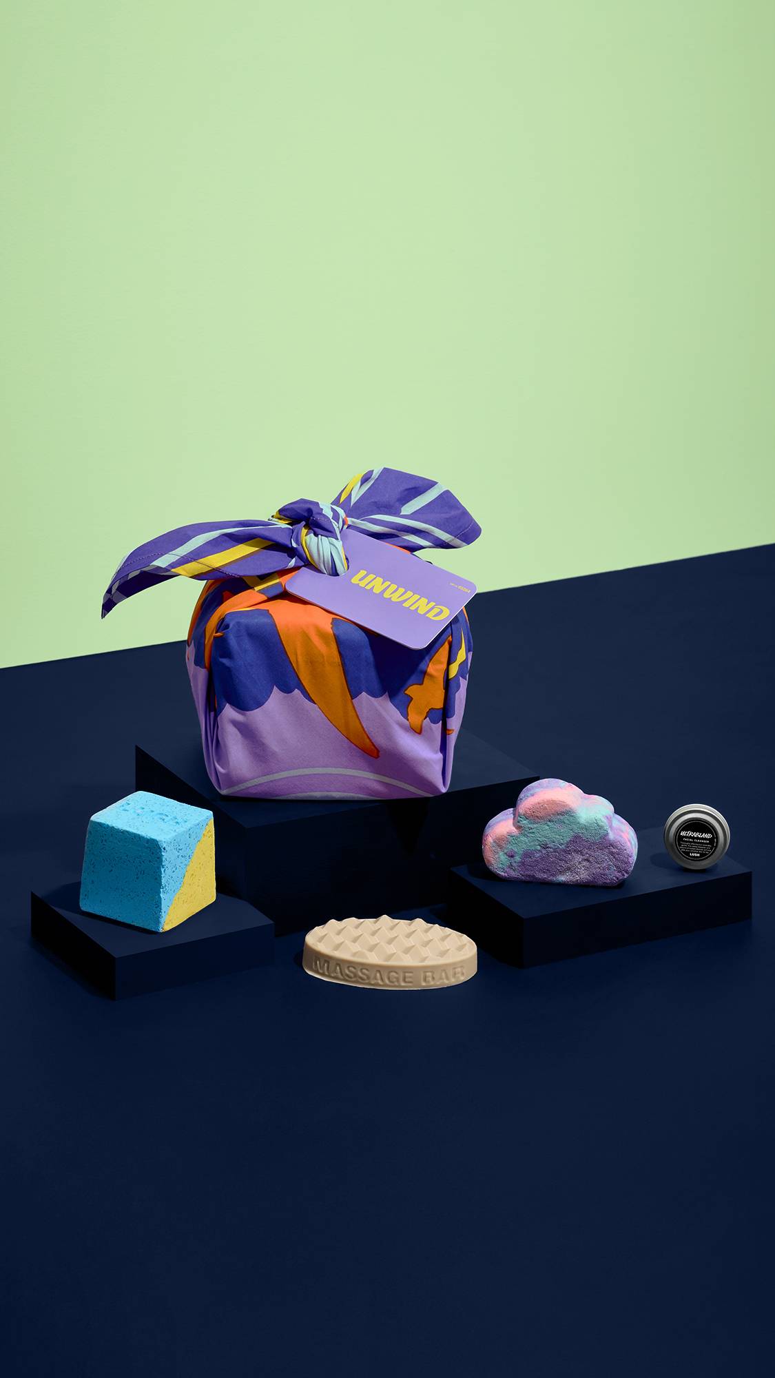 A fabric-wrapped "UNWIND" gift set sits on a dark blue pedestal surrounded by a blue and yellow bath bomb, a waffle-patterned massage bar, a cloud bubble bar, and a small tin, set against a dark blue table and pale green wall.