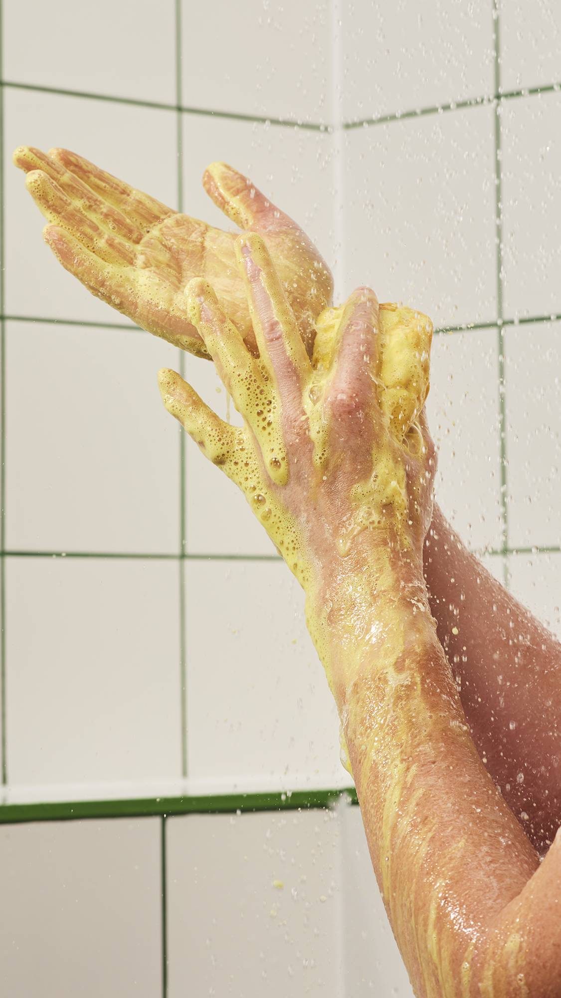 Hands lather a textured yellow cube soap bar under streaming shower water, working up rich, thick yellow foam across hands and arms against a white tiled wall background.