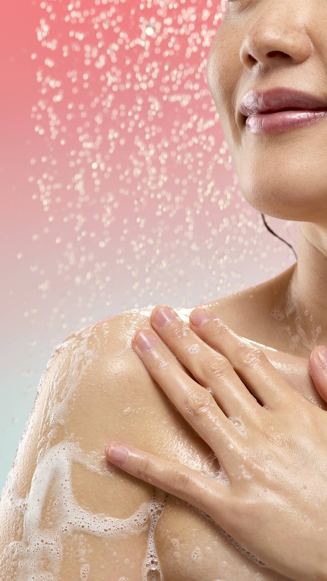 A close-up of a person applying foamy white lather from Witchetty Grub soap to their shoulder while standing under a spray of water. The background features falling droplets against a soft pink-to-blue gradient.