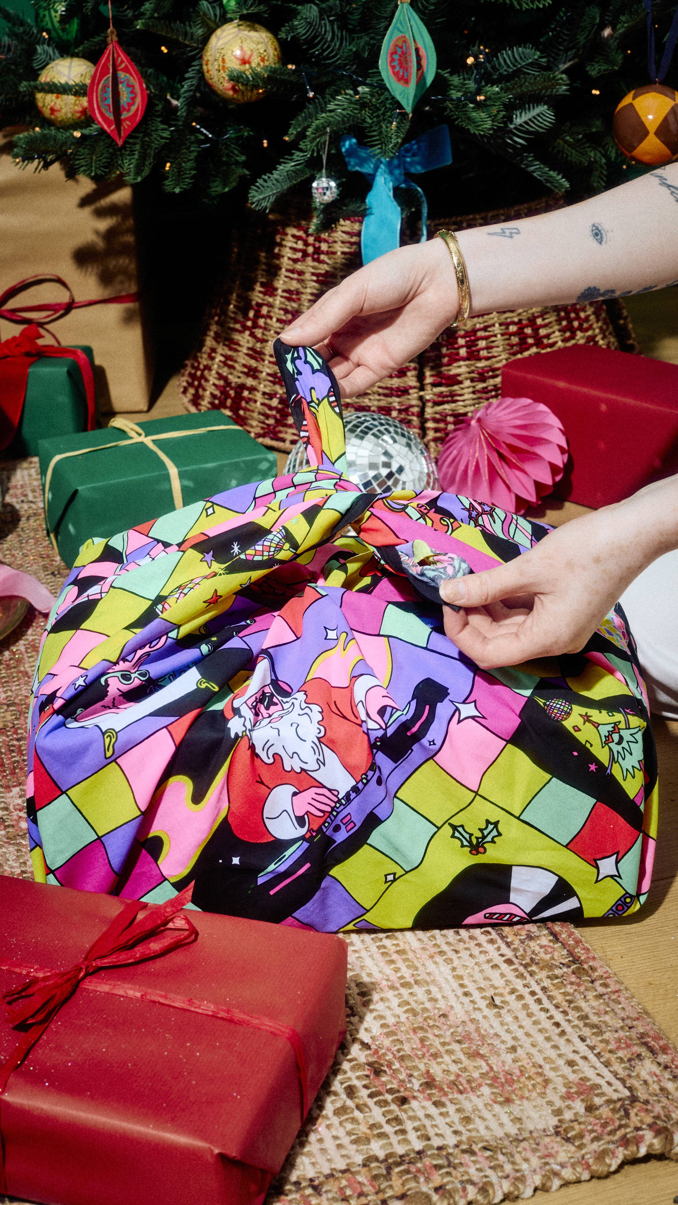 Hands untie the top knot of a colourful, festive disco-patterned Lush fabric Knot Wrap, revealing a wrapped gift beneath a decorated Christmas tree with other gifts nearby.