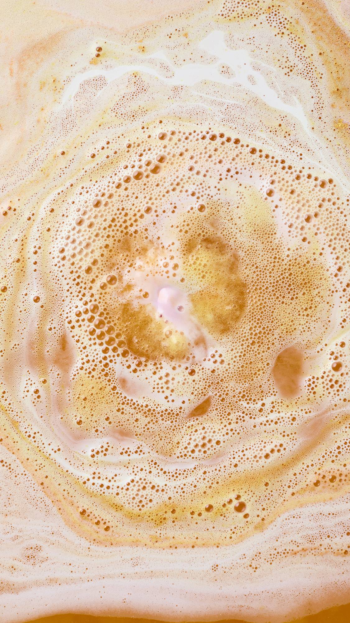 A dissolving Lush "Yog Nog" bath bomb creates swirling rings of warm golden-yellow foam and frothy peach-tinted bathwater, with a small core of soft pink fizz at the very centre.