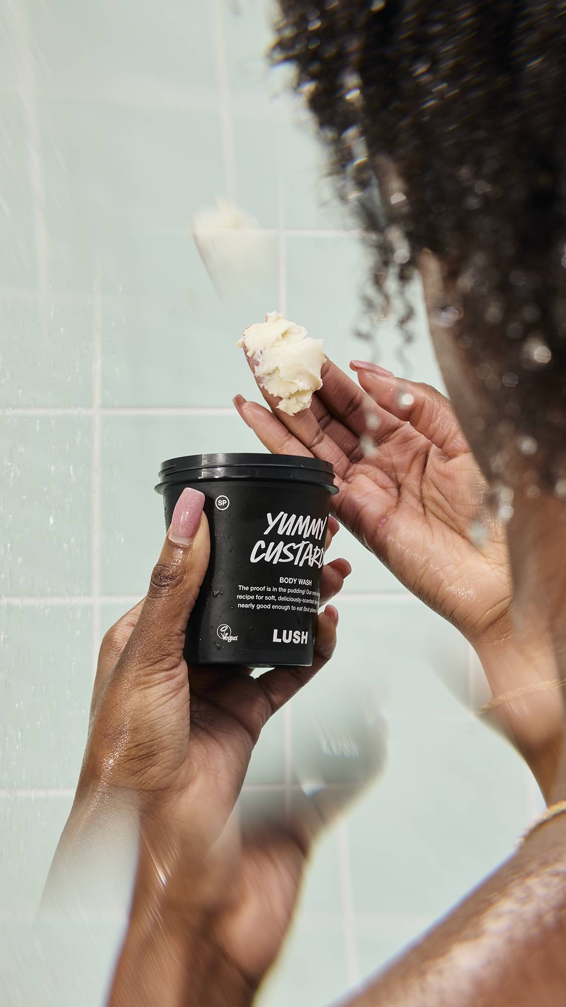 A person scoops a thick dollop of the creamy, yellow "Yummy Custard" body wash from a black pot in the shower. The scene includes falling water droplets and a mint green tiled background, highlighting the product's rich texture.