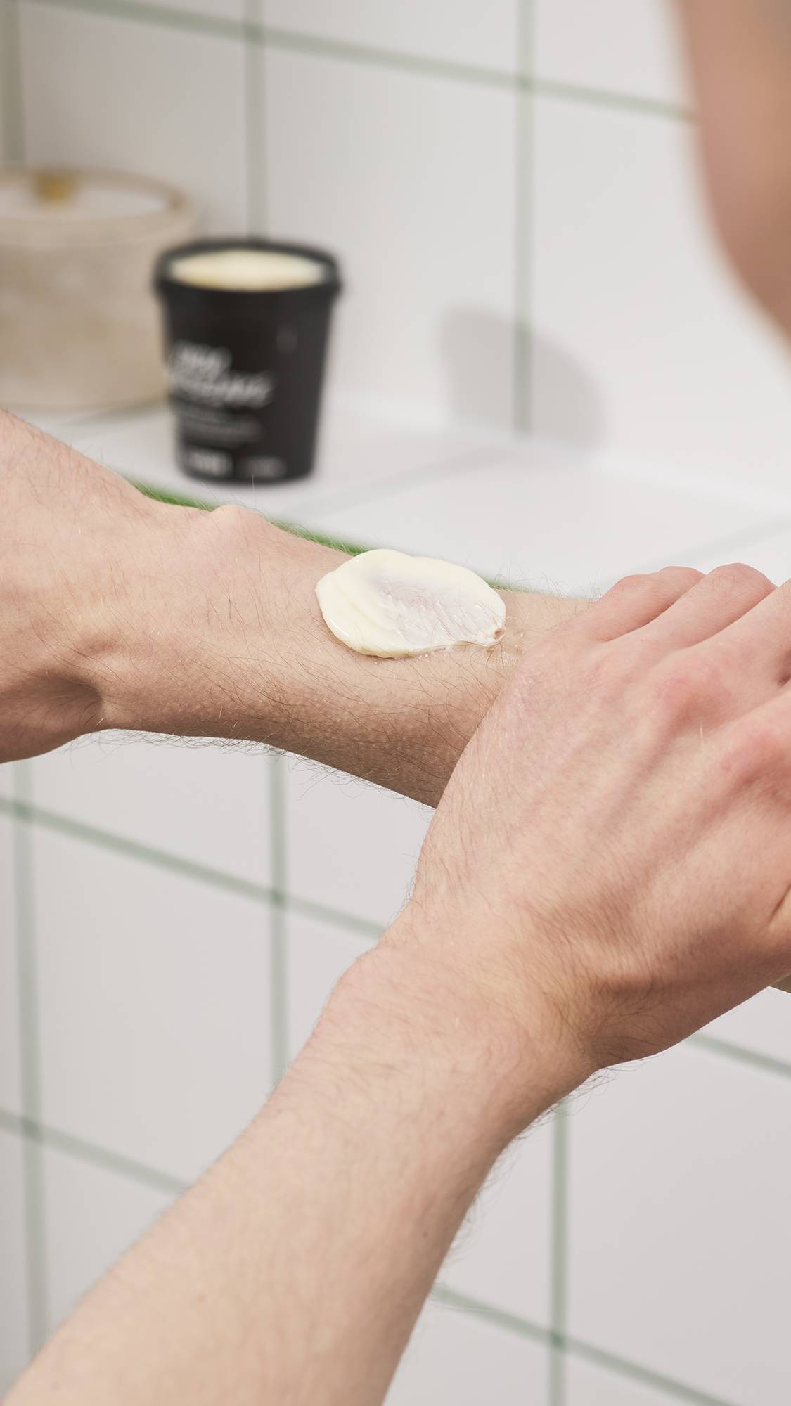A dollop of thick, off-white, Lush "Yuzu Cheesecake" body butter sits on a forearm as a hand begins to apply it, with an open product tub in the background against white tiled bathroom walls.