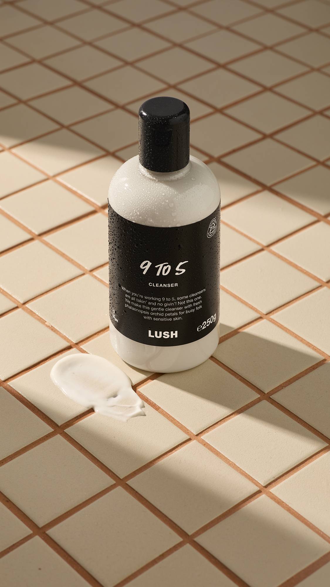 A white bottle of 9 to 5 cleanser with a black label sits on tan tiles, with a dollop of product next to it.
