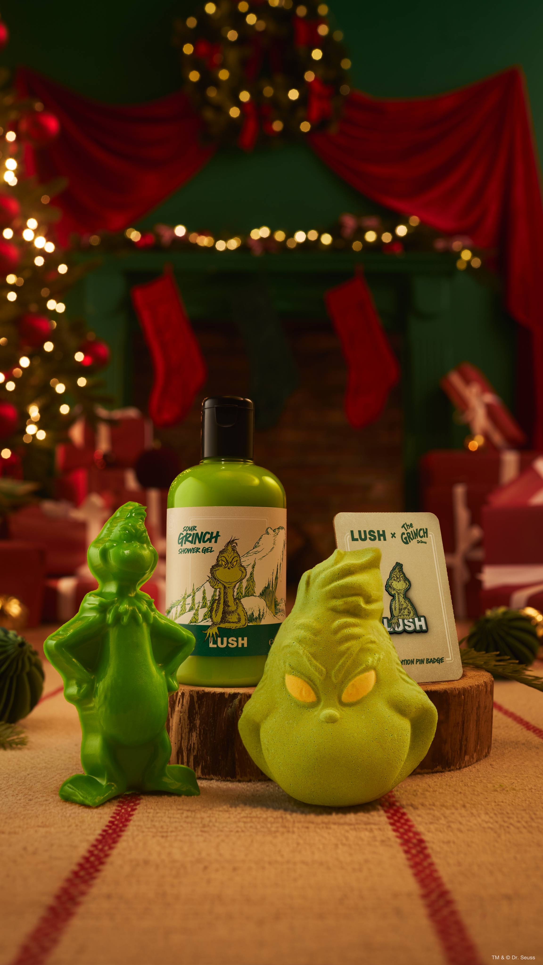 A collection of Lush x The Grinch products, including a green shower gel, a green character-shaped bath bomb, a shower gummy figure, and a pin badge, displayed on a wooden log slice with a blurred festive Christmas background.