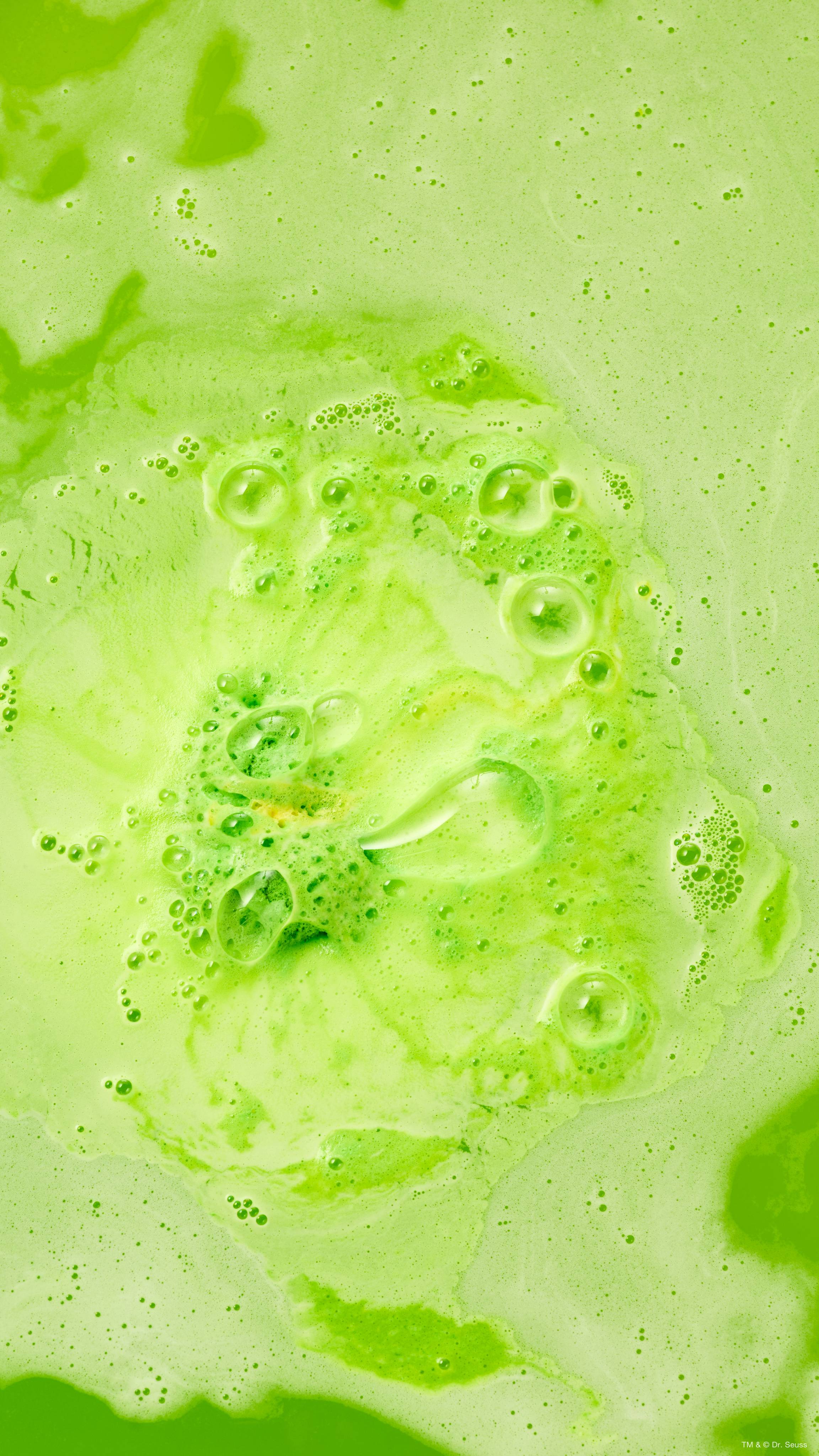 A close-up, aerial shot of a dissolving green "Grinch" bath bomb creating swirling, bright lime-green foam and frothy bubbles across vivid green bathwater.  