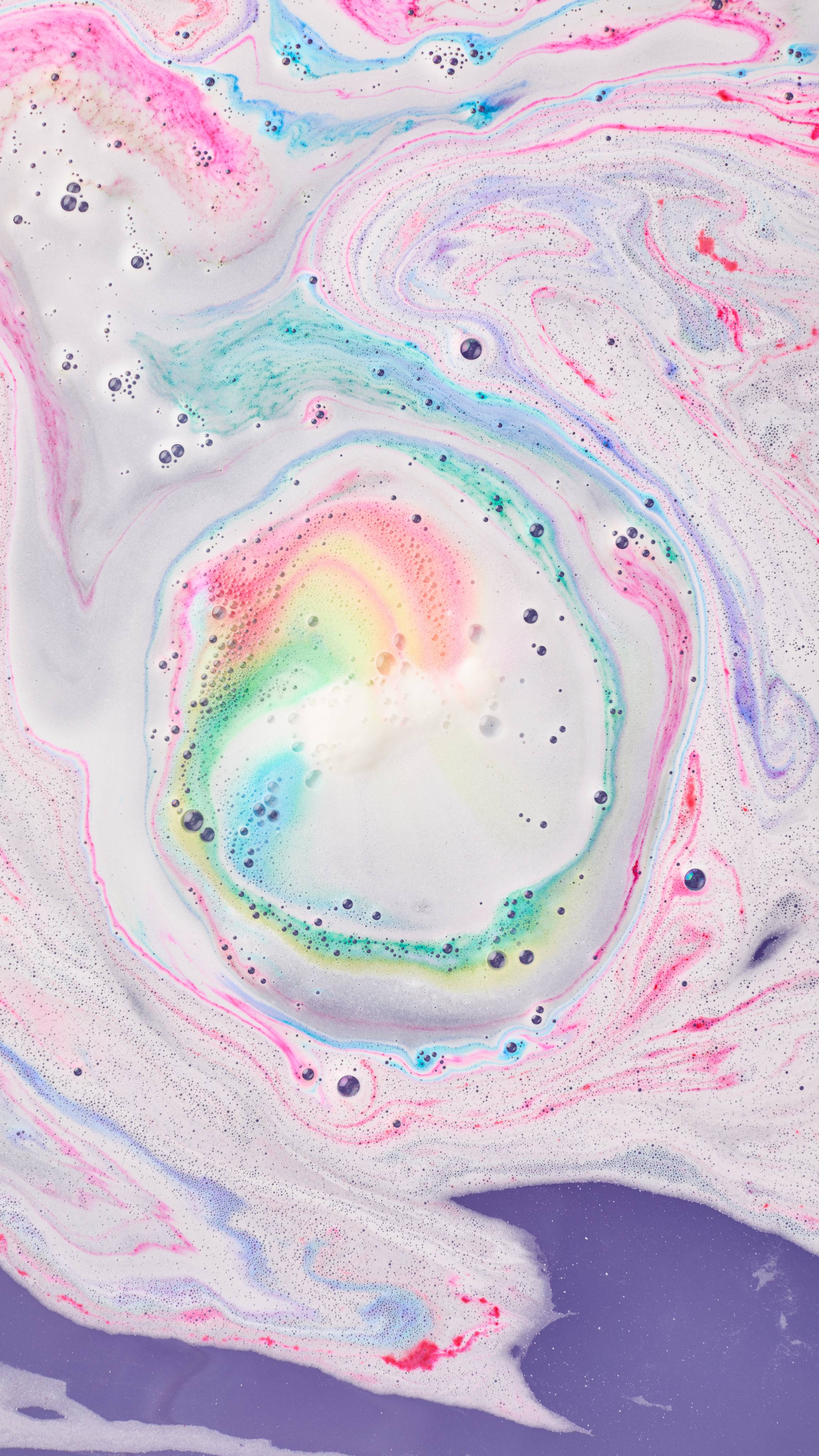 An aerial view of swirling streams of rainbow bath foam in pastel pink, yellow, teal, and blue creates a colourful spiral at the centre of soft white and deep purple water.