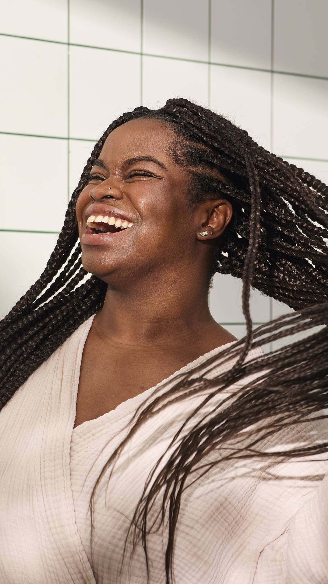 A person with long dark braids laughs joyfully while tossing their hair, wearing a light pink muslin robe. They stand against a background of white square tiles with green grout.
